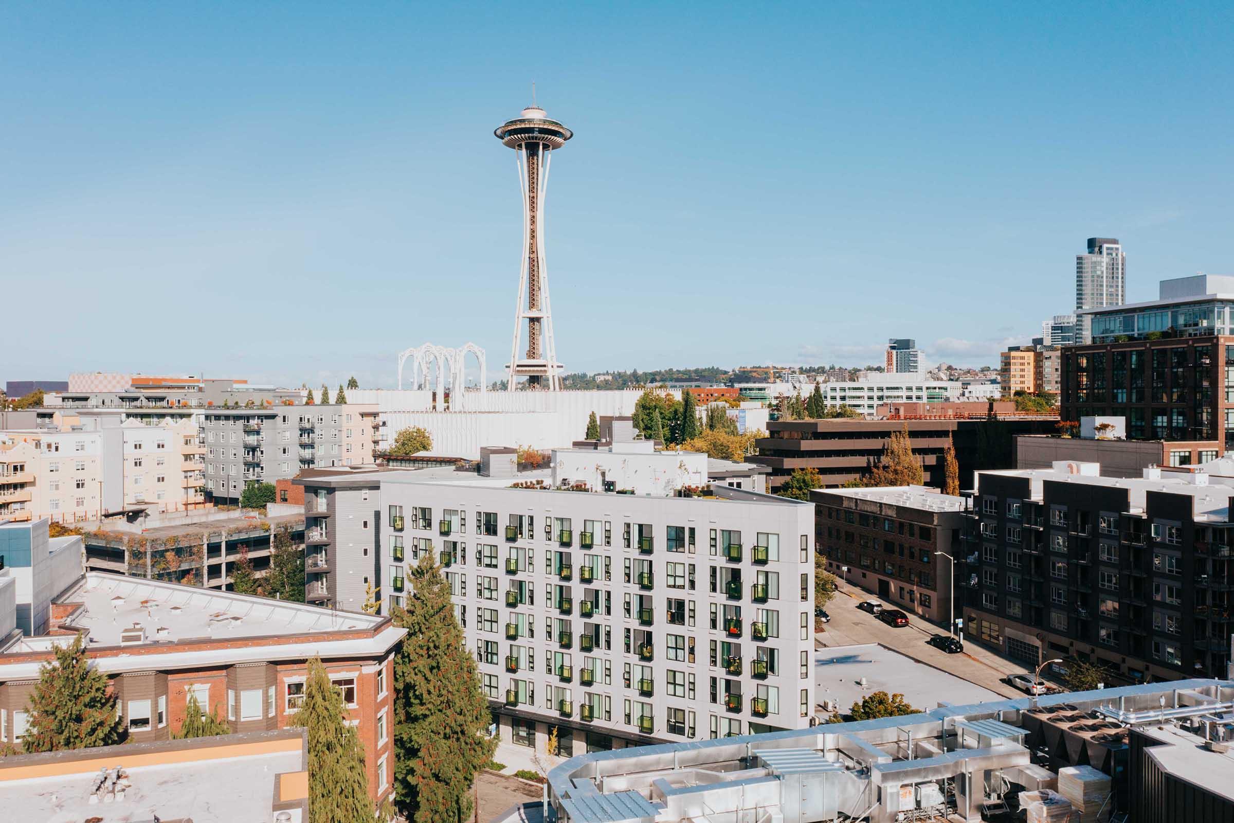 A panoramic view of a cityscape featuring the Space Needle in Seattle. In the foreground, there are modern buildings with greenery, while the background showcases a clear blue sky and urban structures. The scene captures a vibrant mix of residential and commercial architecture.