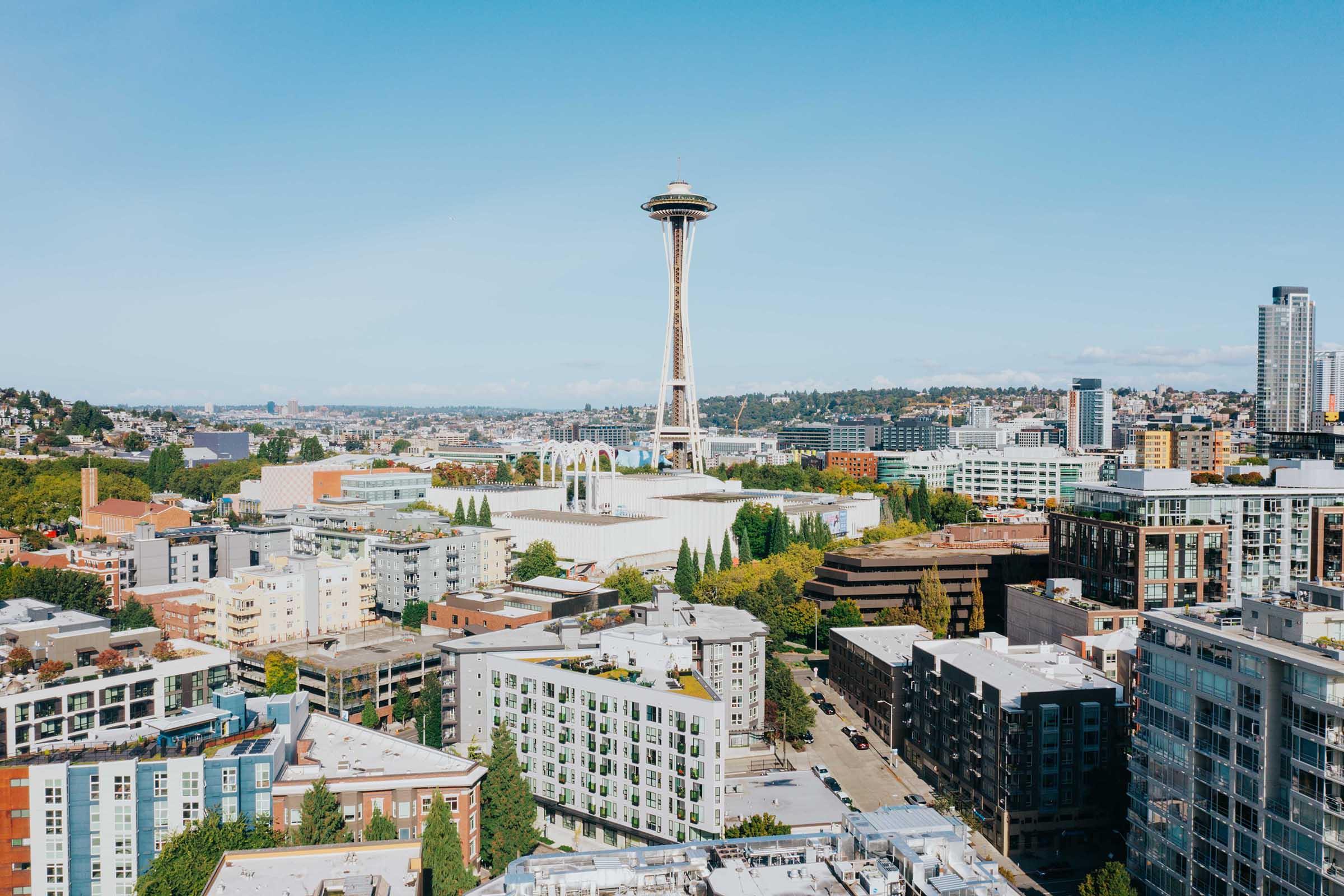 A panoramic view of Seattle featuring the iconic Space Needle in the background, surrounded by modern buildings and green trees. The scene is set against a clear blue sky, showcasing the urban landscape of the city.