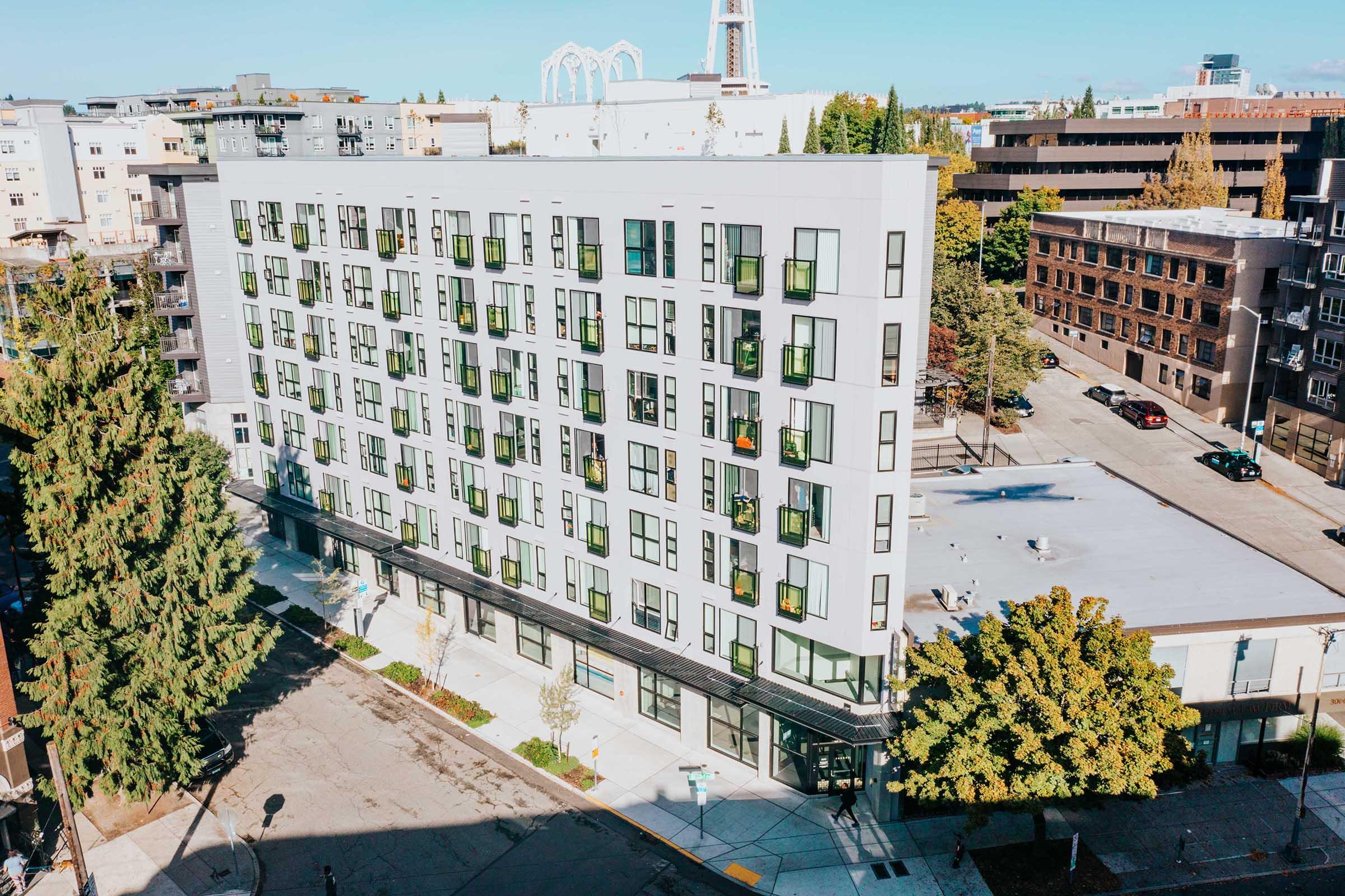 Modern multi-story residential building with numerous windows, surrounded by trees and urban infrastructure. Clear blue sky overhead, and a hint of nearby structures in the background. Street view showcasing a well-maintained sidewalk and landscaping.