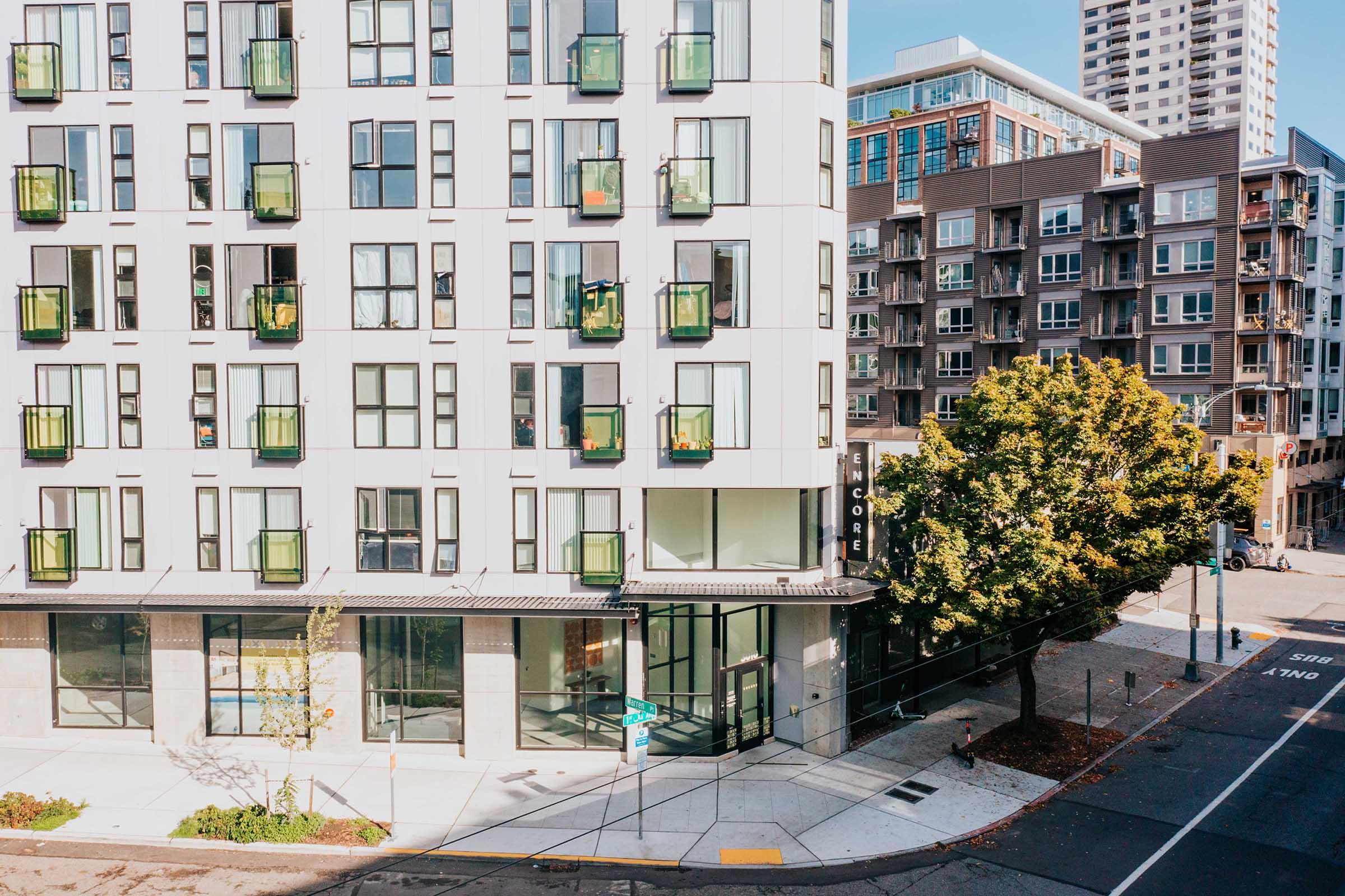 A modern multi-story residential building with balconies featuring green planters. The ground level has large glass windows, and there are trees and sidewalks nearby. In the background, another apartment building is visible, showcasing a mix of architectural styles in an urban setting.