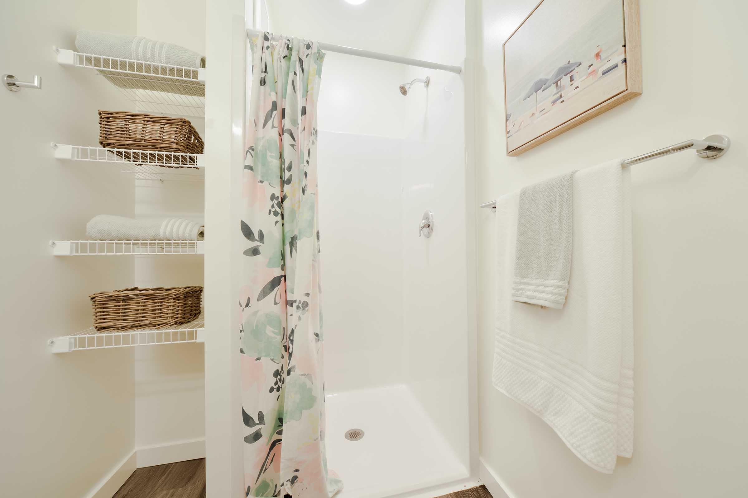 A bright and modern bathroom featuring a walk-in shower with a floral curtain. There are neatly arranged towels on a rack and woven baskets on open shelves, creating a clean and organized look. A framed artwork hangs on the wall, adding a decorative touch.