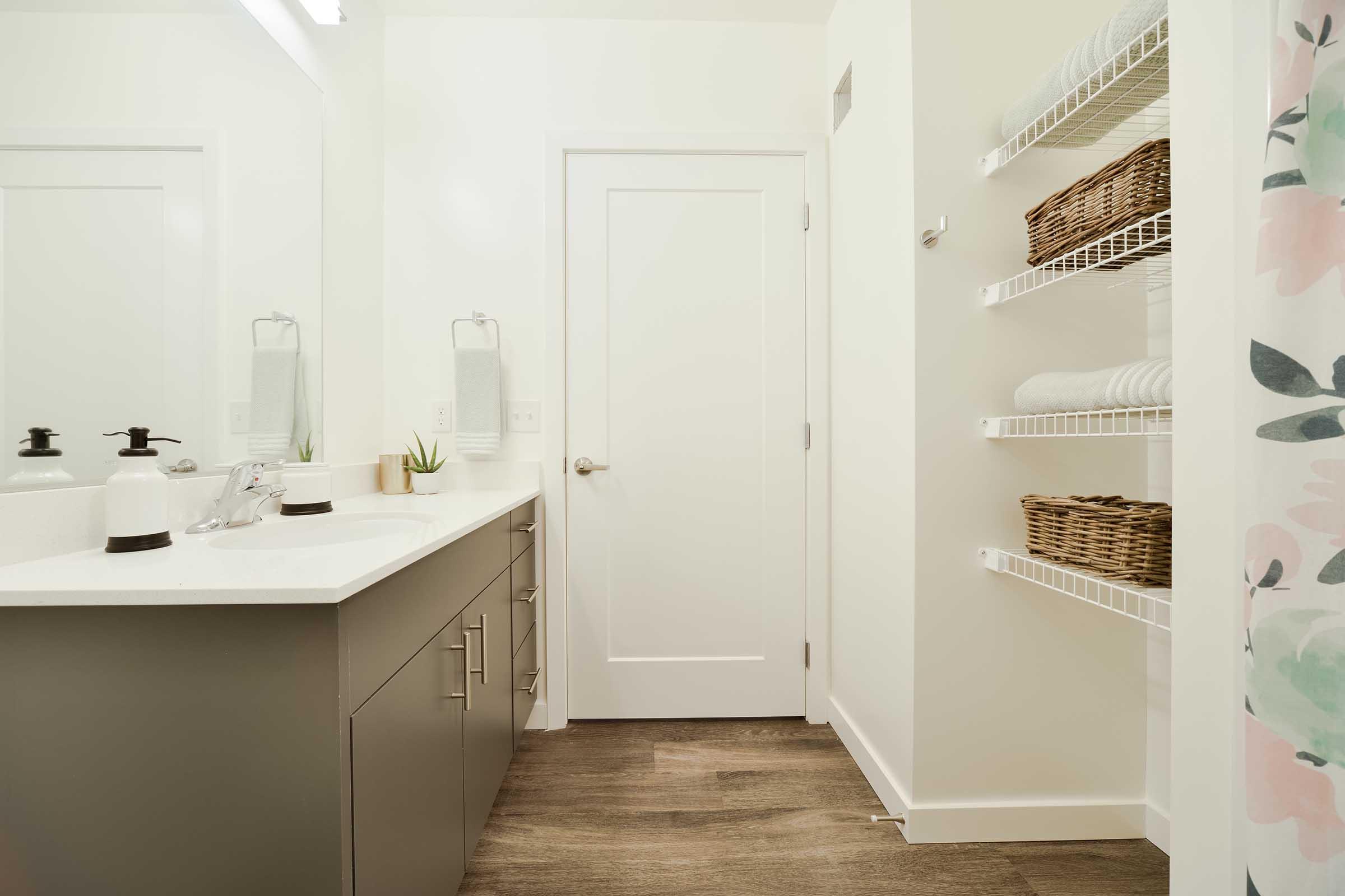 A modern bathroom featuring a white countertop with a sink and sleek dark cabinetry. The walls are light-colored, and a door leads to an adjoining space. Shelving holds woven baskets and neatly stacked towels. A shower curtain with a floral design is visible, enhancing the fresh and clean aesthetic.