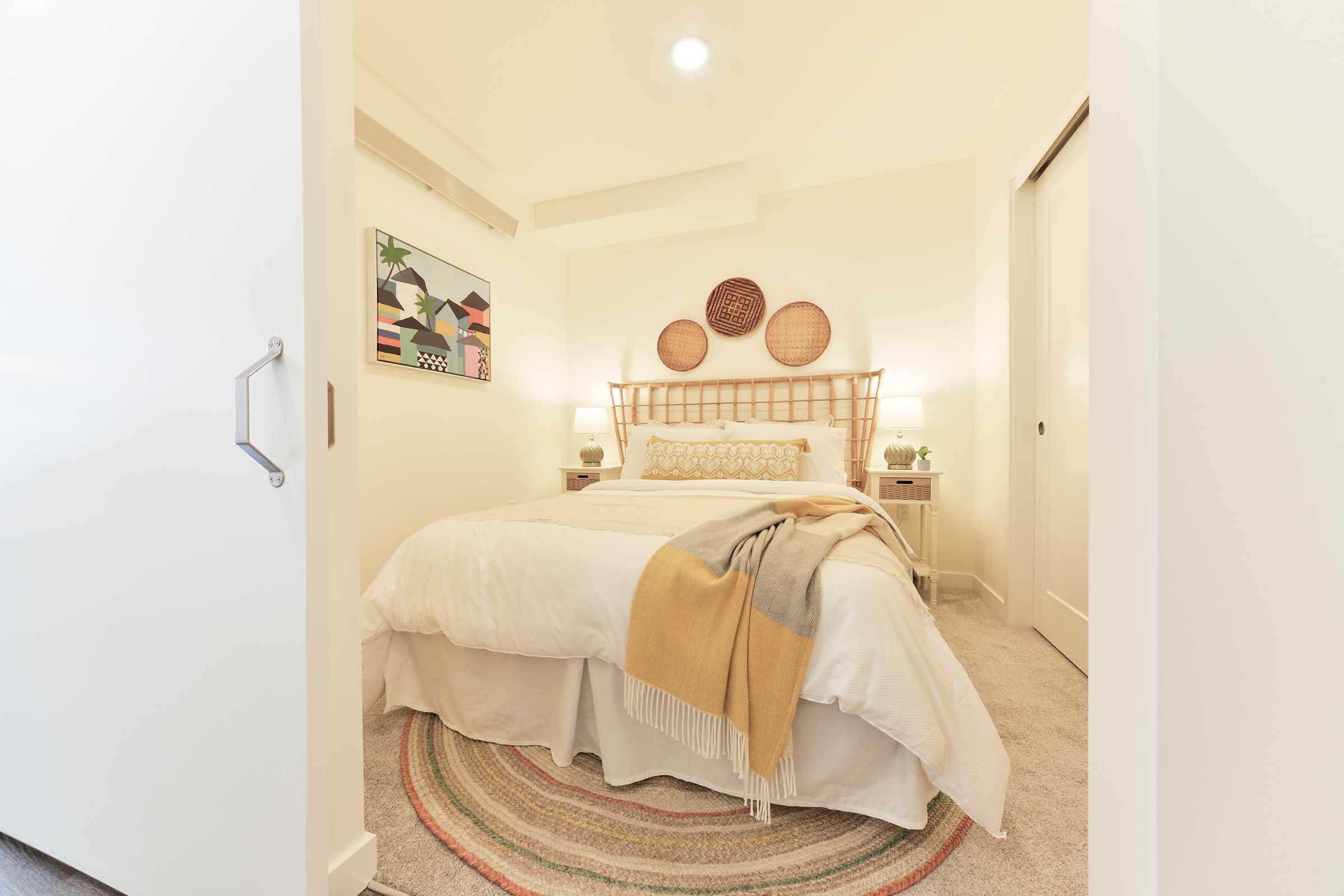 A cozy bedroom featuring a bed with a light-colored duvet and decorative pillows. The walls are adorned with woven wall art and a colorful painting. Soft lighting from bedside lamps complements a round rug on the carpeted floor, creating a warm and inviting atmosphere.