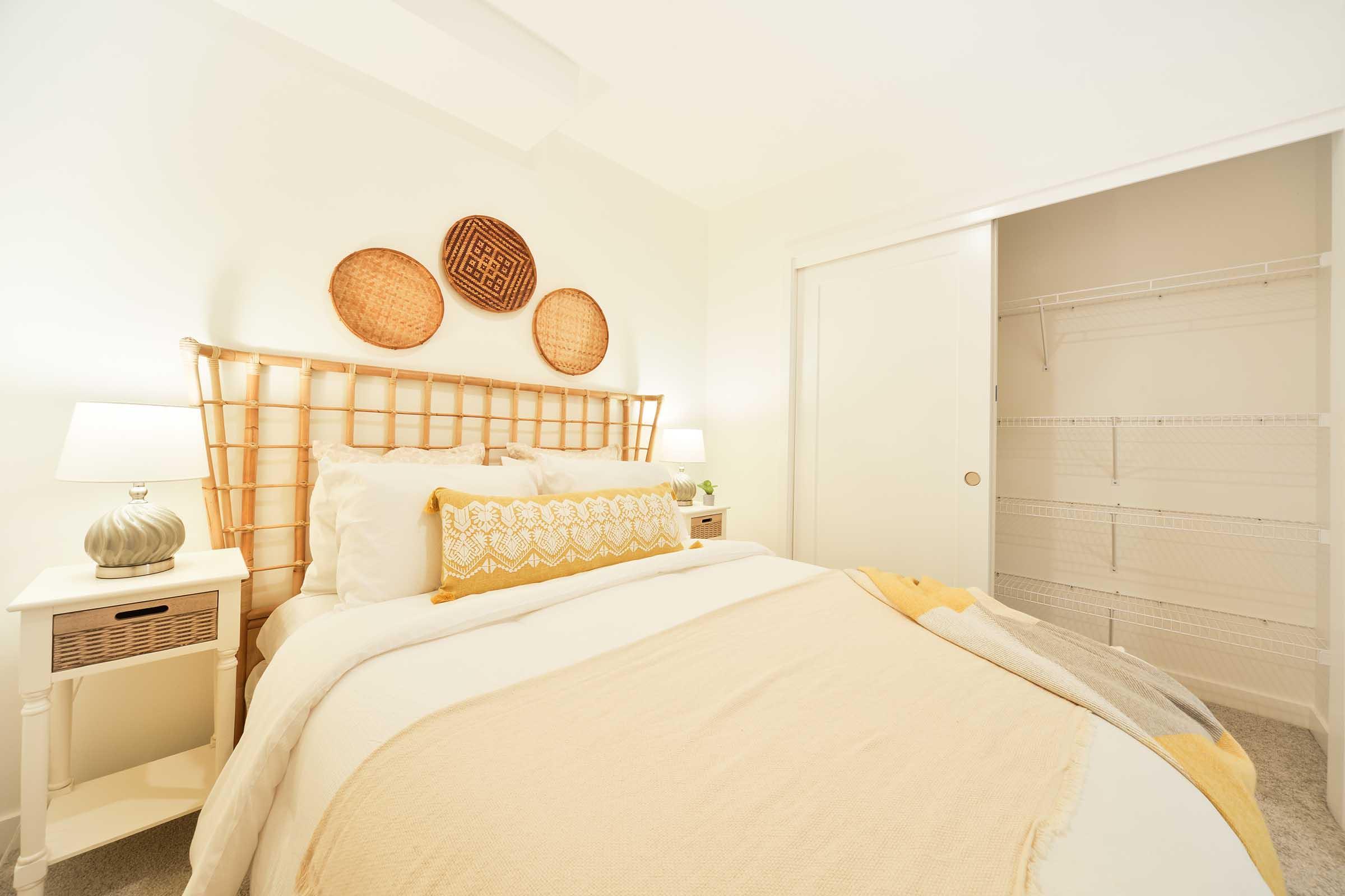 A bright and cozy bedroom featuring a queen-sized bed with a light beige and yellow throw blanket. There are two bedside tables with lamps, woven wall decor above the headboard, and a spacious closet with sliding doors, all set against a soft white backdrop.