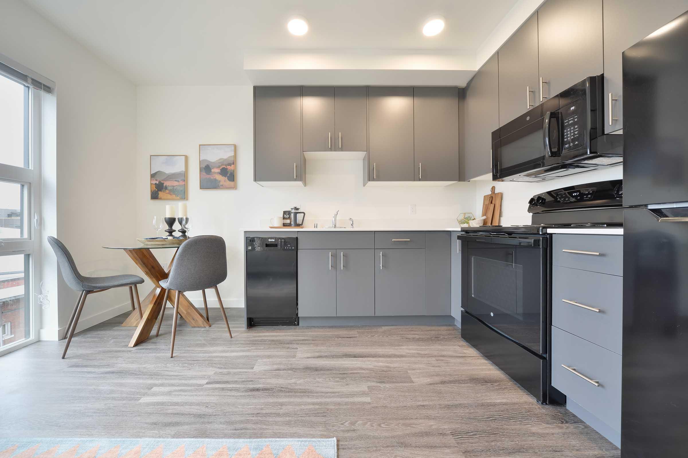 Modern kitchen featuring gray cabinets and black appliances. A small dining table with two gray chairs is positioned nearby. The space has large windows allowing natural light, and there are decorative items on the table and walls, creating a stylish and inviting atmosphere.