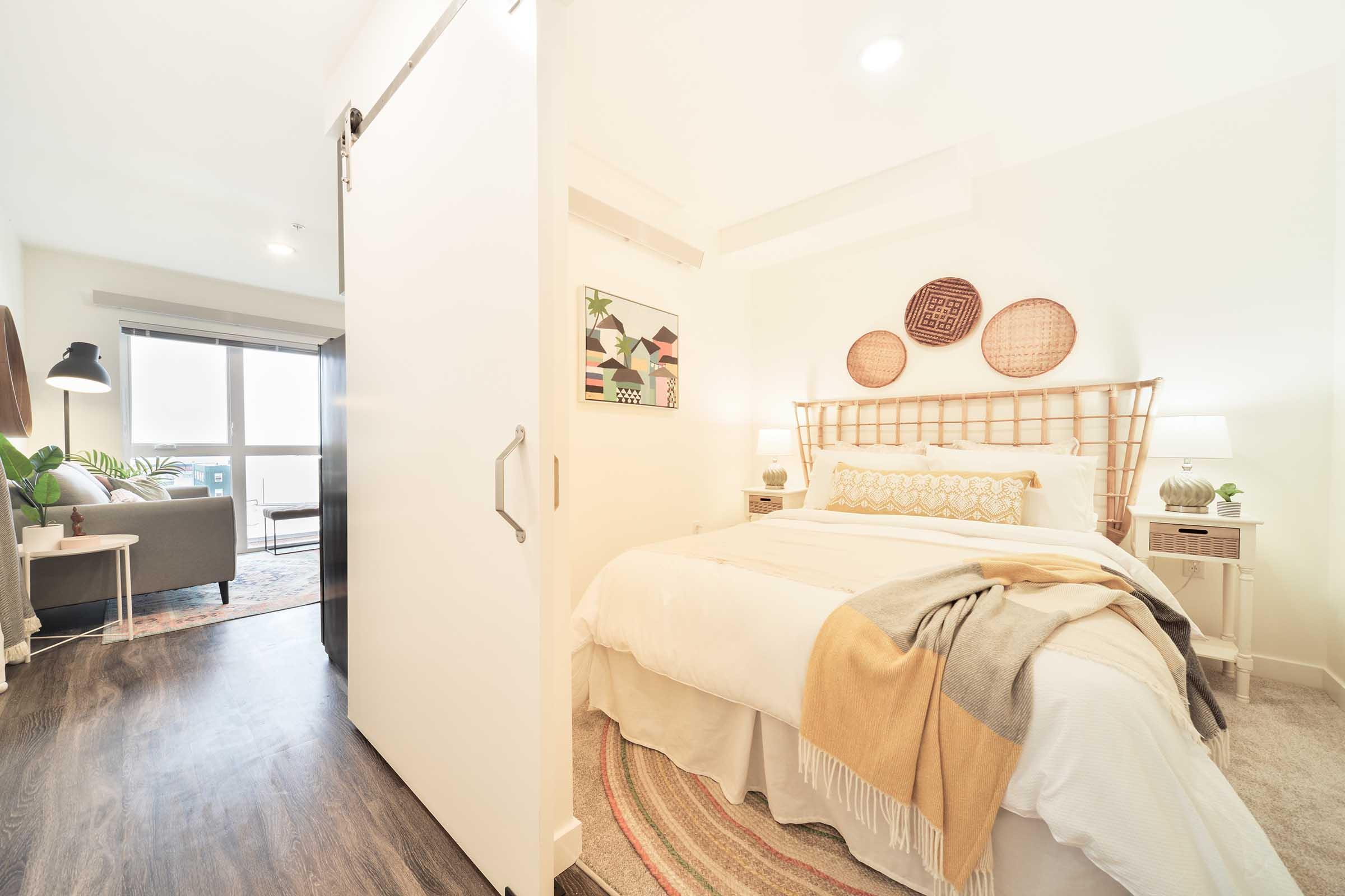 A cozy bedroom featuring a bed with a light-colored blanket and decorative pillows. There are woven wall hangings, a bedside table with a lamp, and a sliding door leading to a living area with a couch and plants. Natural light fills the space, creating a warm and inviting atmosphere.