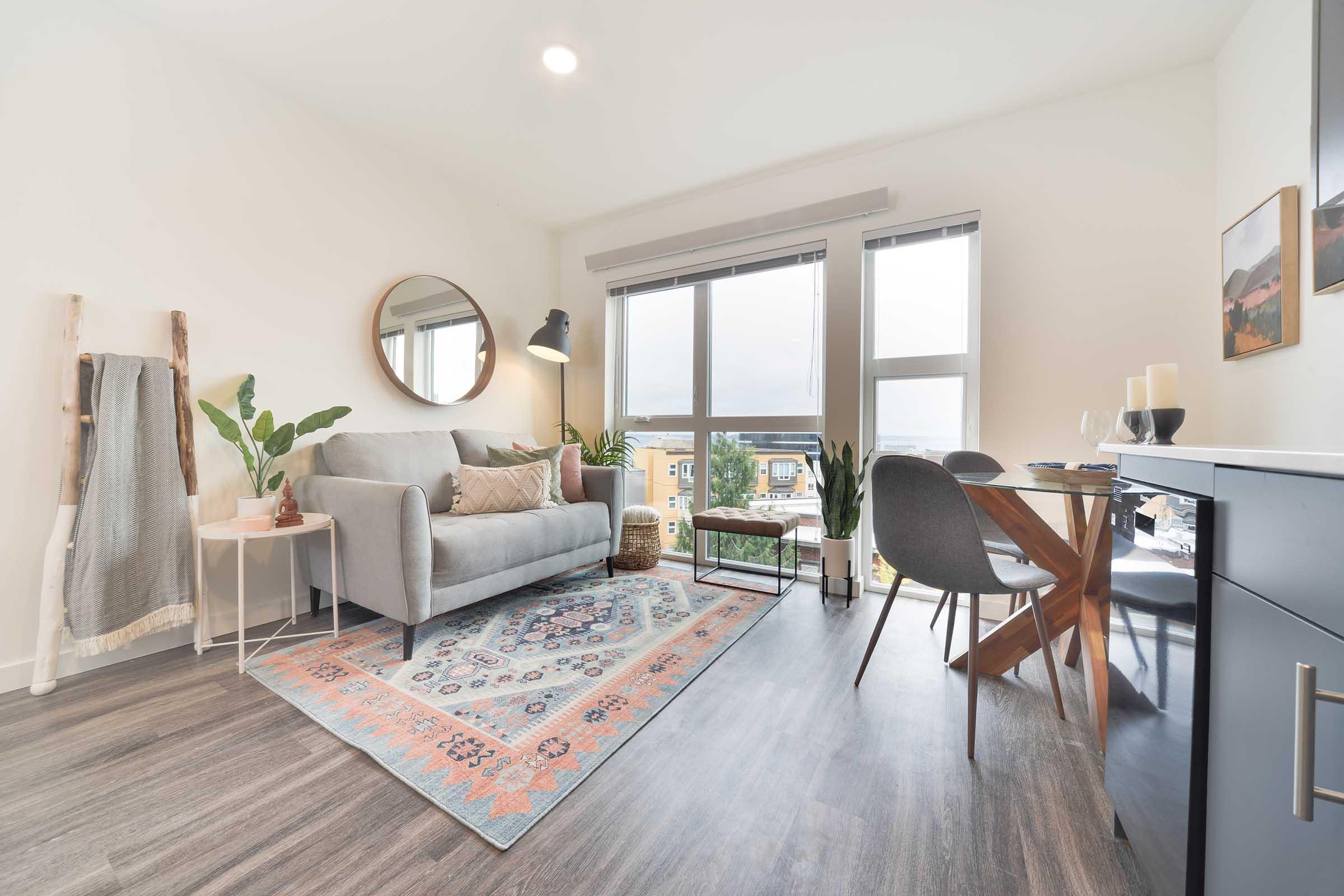 A modern living room with a light gray sofa and a round mirror above it. There's a potted plant on a side table, and a patterned rug on the floor. Large windows provide natural light, with a view outside. A dining area features a wooden table and gray chairs, complemented by a dark storage cabinet.