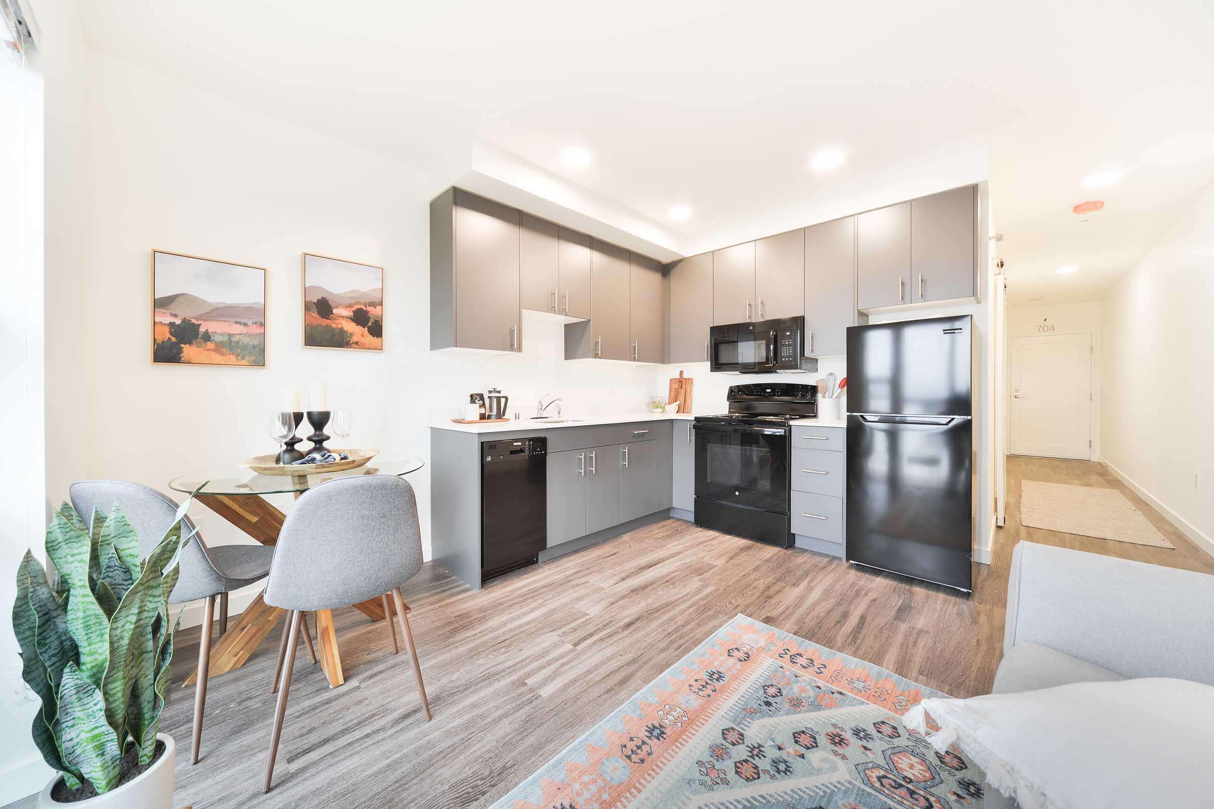 A modern kitchen with dark cabinets, black appliances, and a small dining area featuring a round glass table and gray chairs. Houseplants add a touch of greenery, while artwork adorns the walls. The space has a bright, airy feel with natural light coming in.