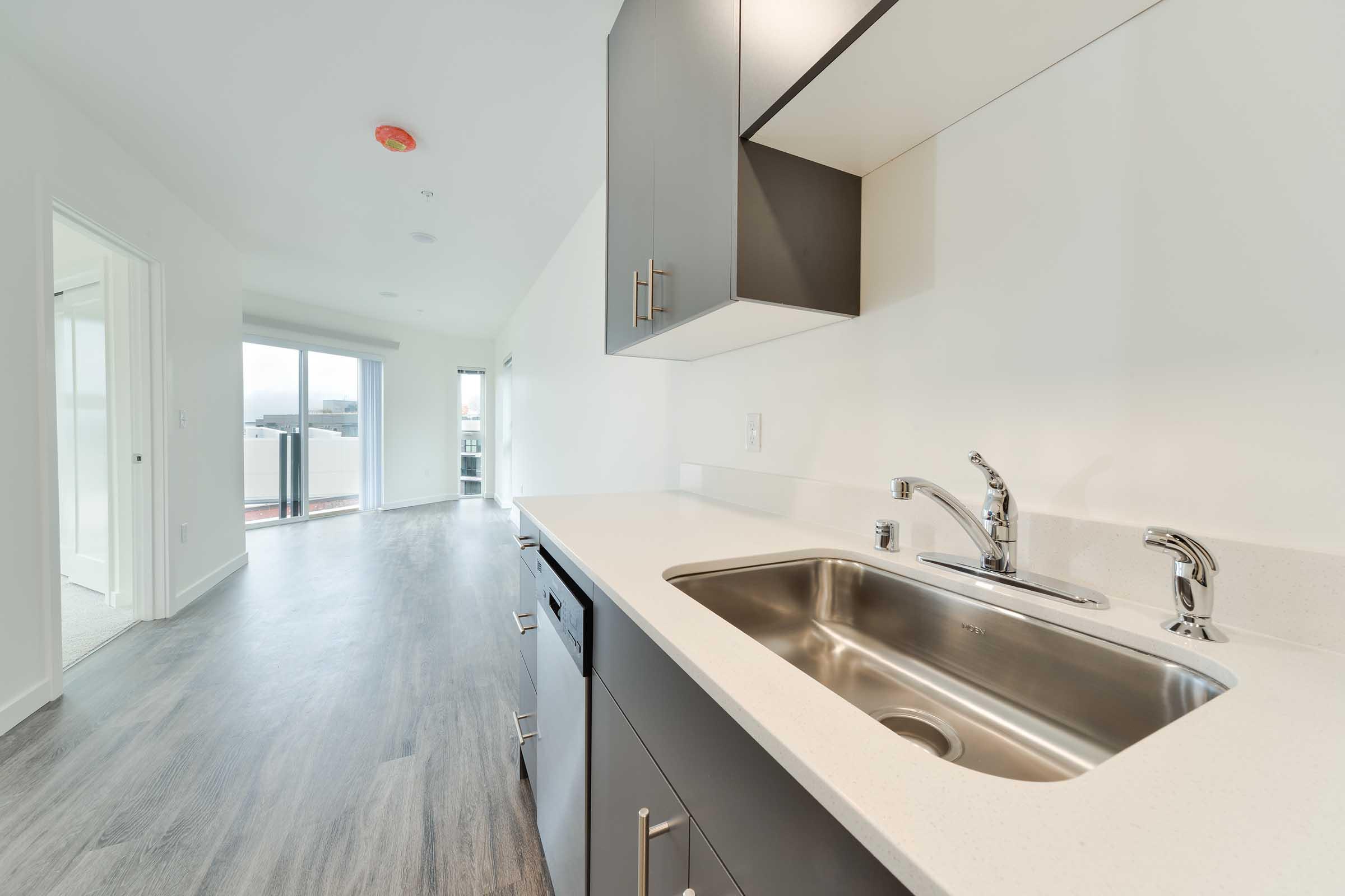 A modern kitchen featuring a stainless steel sink with a minimalist design. The cabinetry is dark gray, and there are silver faucets. The image also shows an open layout leading to a bright living space with large windows and a balcony in the background, illuminated by natural light.