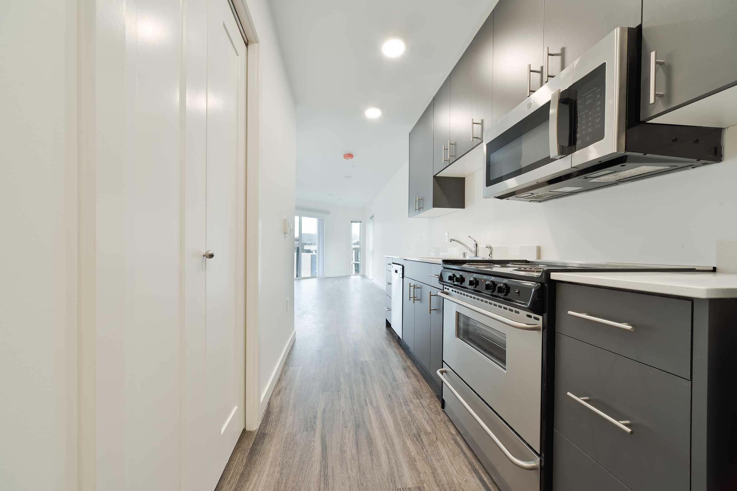 Modern kitchen featuring stainless steel appliances, including a gas stove and microwave. The cabinetry is dark grey, and the countertops are light. The space has a sleek, clean design with bright overhead lighting and a doorway leading to a light-filled living area.