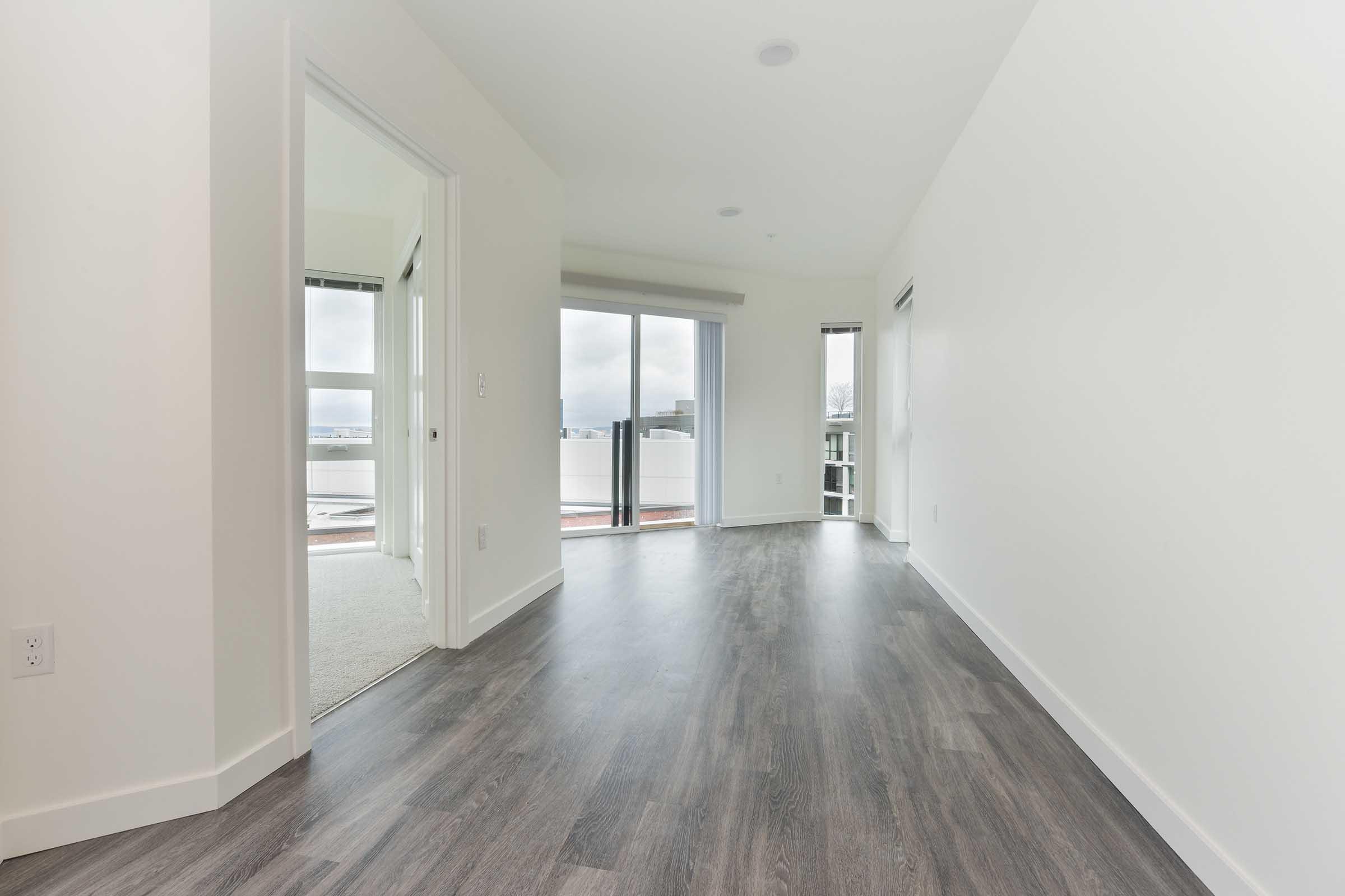 A spacious, modern room with light-colored walls and dark wood-like flooring. There are two doorways leading to other areas, and large windows allowing natural light to enter. The view outside includes a cloudy sky, and there is a balcony visible through the glass doors.