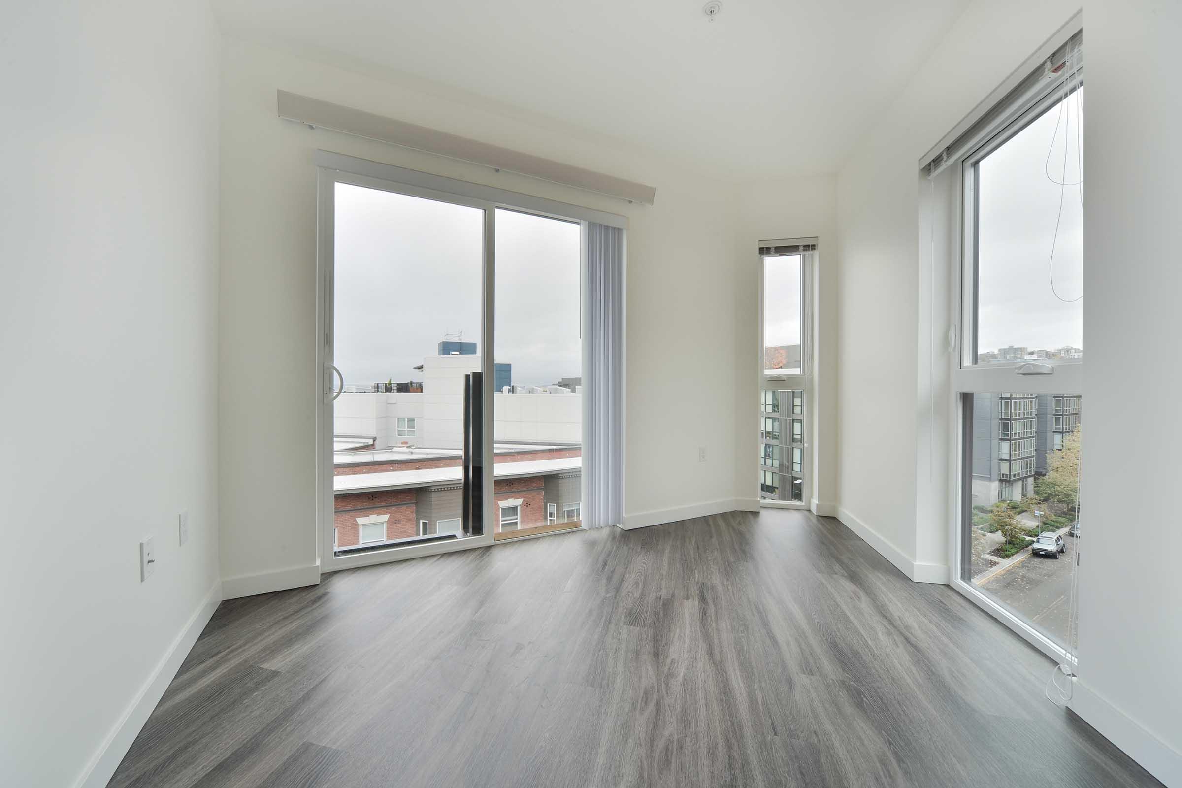 A bright, empty room with large windows showcasing a city view. The floor has a modern gray finish, and the walls are painted white. Natural light fills the space, highlighting the minimalist design. The room features sliding glass doors leading to an outdoor area.