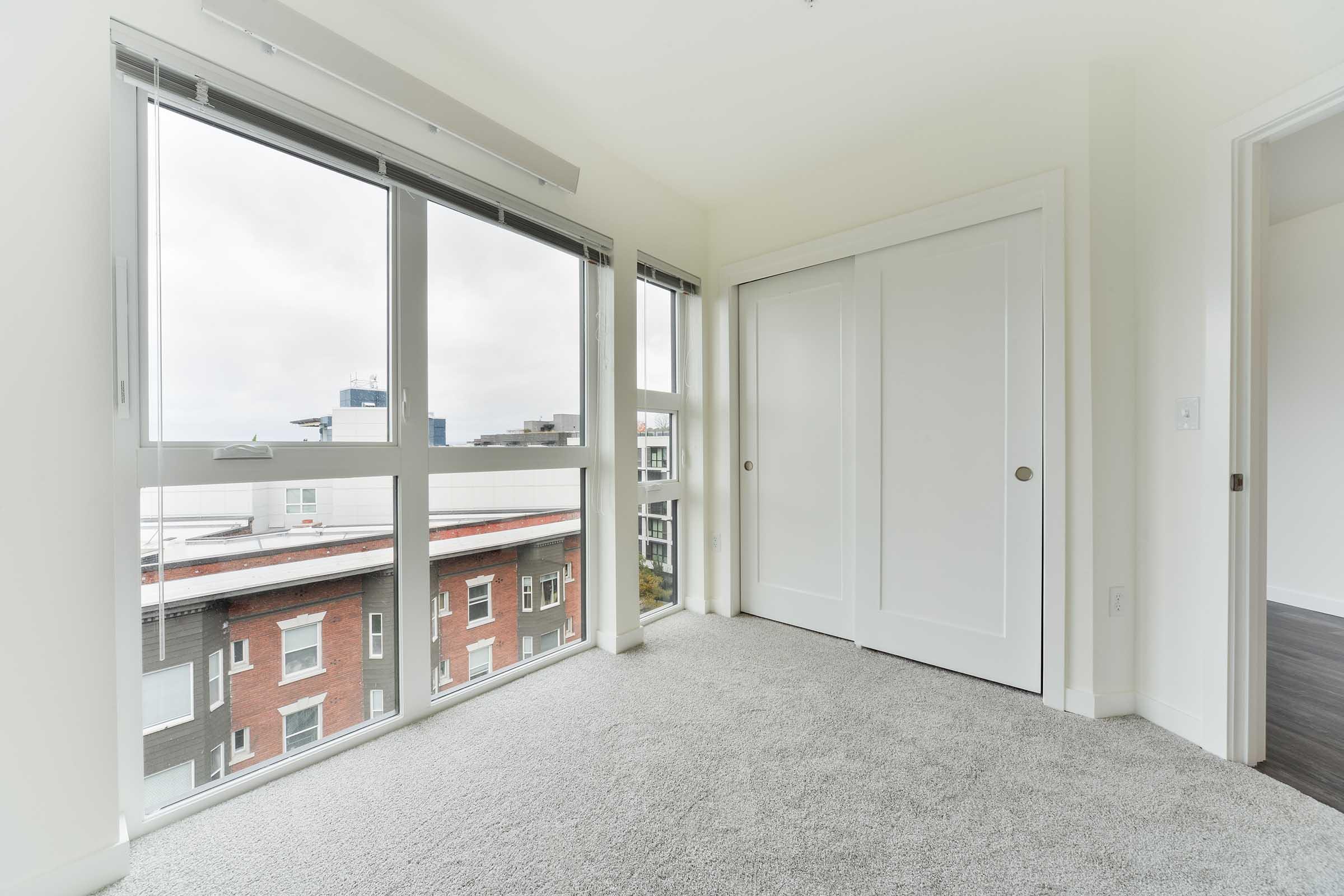 A bright, empty bedroom featuring large windows that offer a view of a cityscape. The room has light-colored walls and carpet, along with a closet that has double doors. Soft natural light fills the space, creating a clean and modern atmosphere.