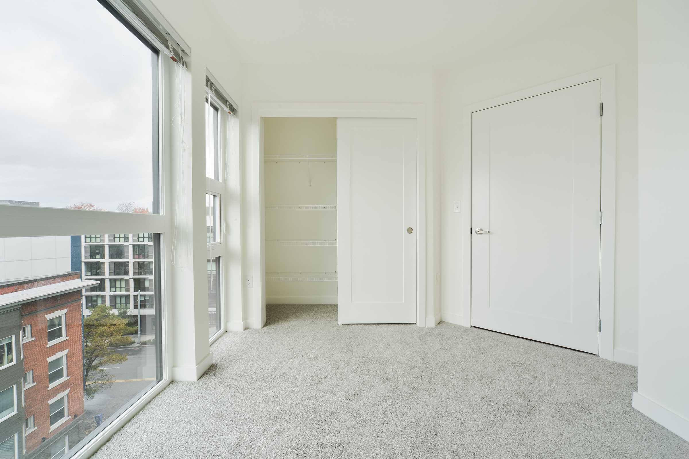 A bright, empty room with light gray carpet. It features large windows allowing natural light, a closet with a sliding door, and a white door leading to another area. The walls are painted white, creating a clean and spacious atmosphere. Urban views can be seen through the windows.