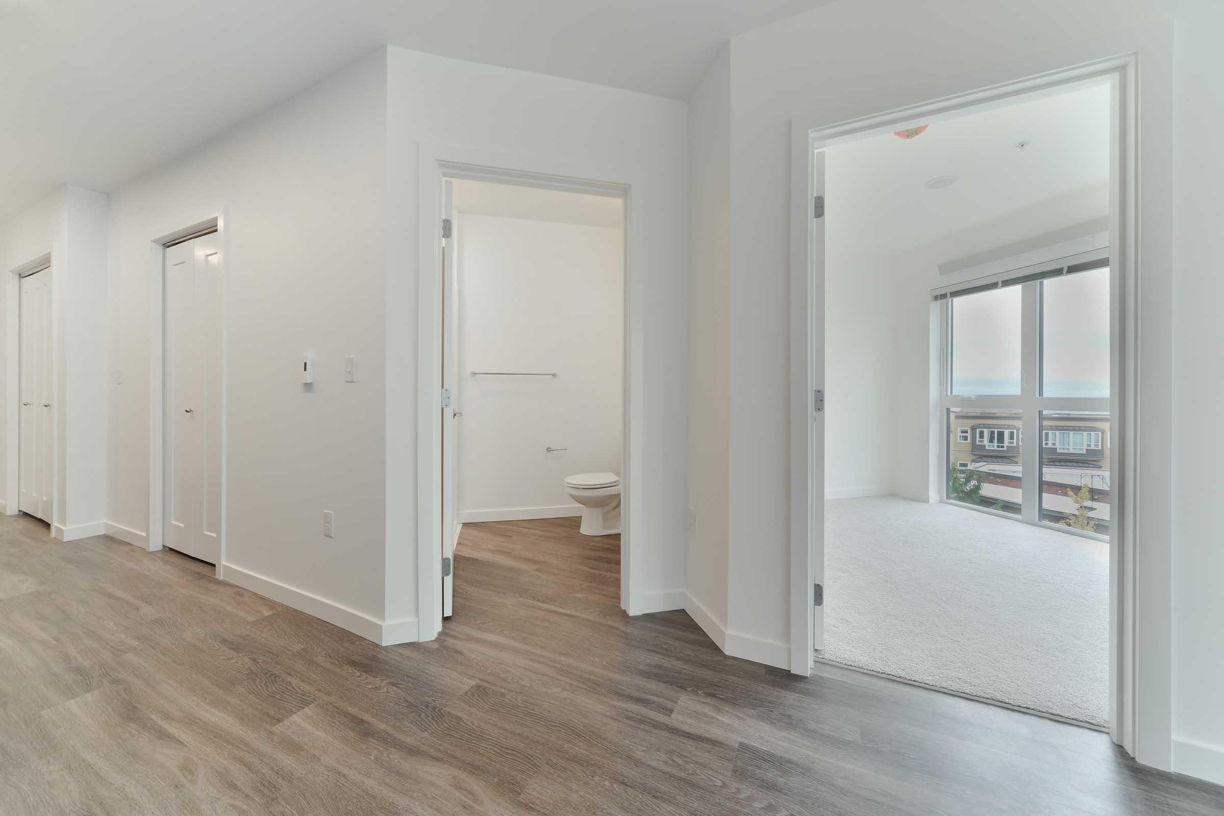 A modern, well-lit hallway featuring multiple doors, leading to a bathroom on the left with minimal decor. On the right, there's an open doorway revealing a bright, carpeted room with large windows showcasing a view. The flooring is a neutral wood tone, contributing to a contemporary aesthetic.