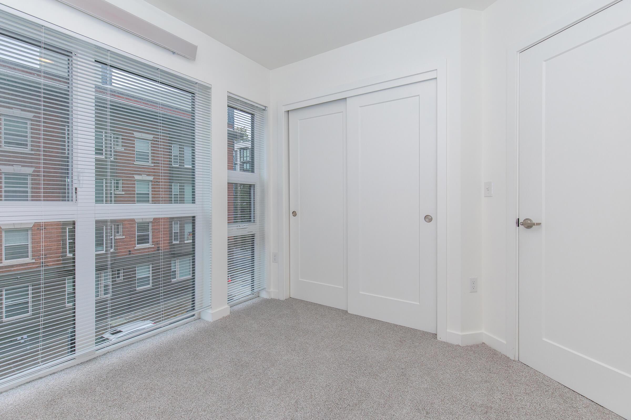 A bright, modern room featuring large windows with blinds, showcasing a view of a building across the street. The room has light-colored walls, plush carpet flooring, and a closet with double doors, creating a clean and spacious feel. A white door leads to another area, completing the minimalist design.