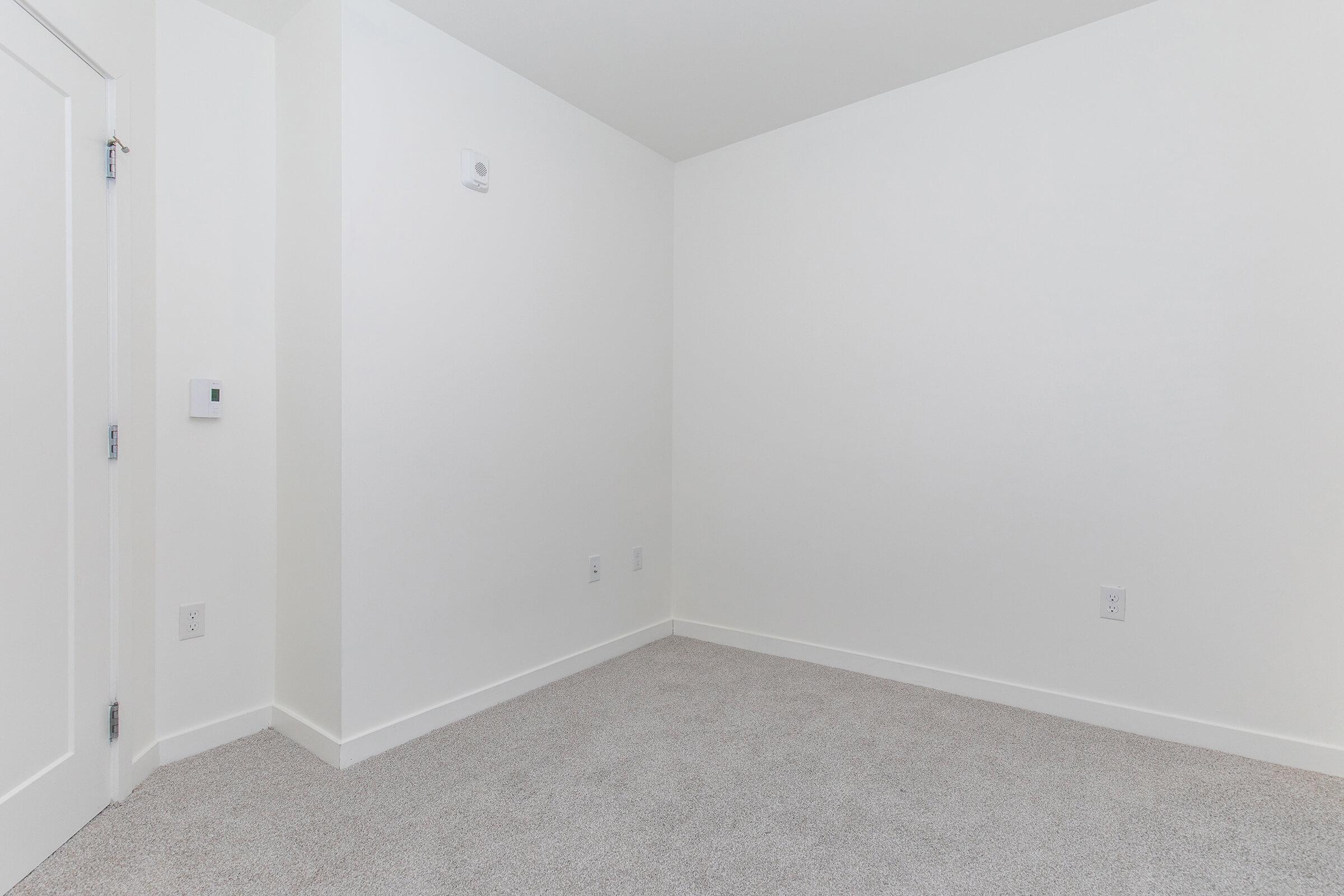 A small, empty room with white walls and light beige carpet. There is a light switch on the left wall and a small white device mounted near the ceiling. The space is well-lit and uncluttered, giving a minimalist appearance.
