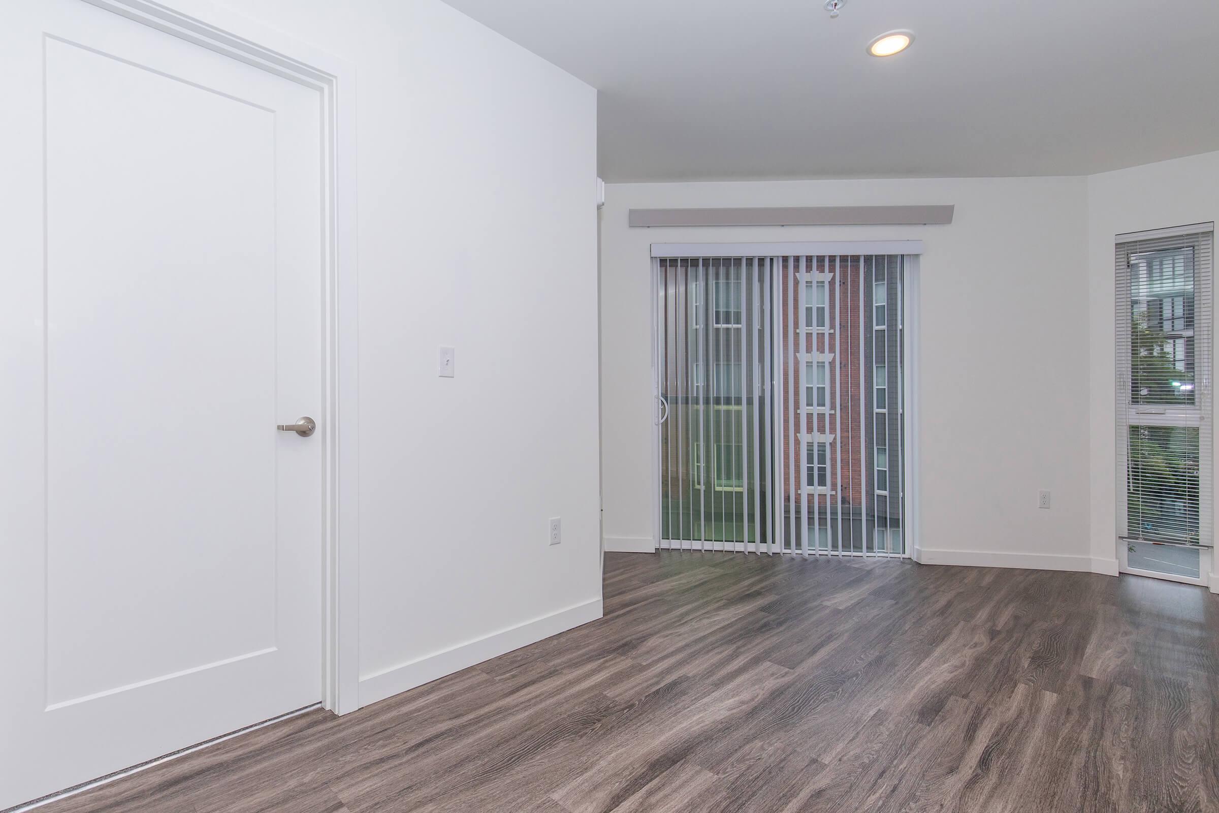 A bright, empty room featuring light-colored walls and a sliding glass door with vertical blinds. The floor is wooden, and there is a plain white door on the left. Natural light streams in through the door, illuminating the spacious area.