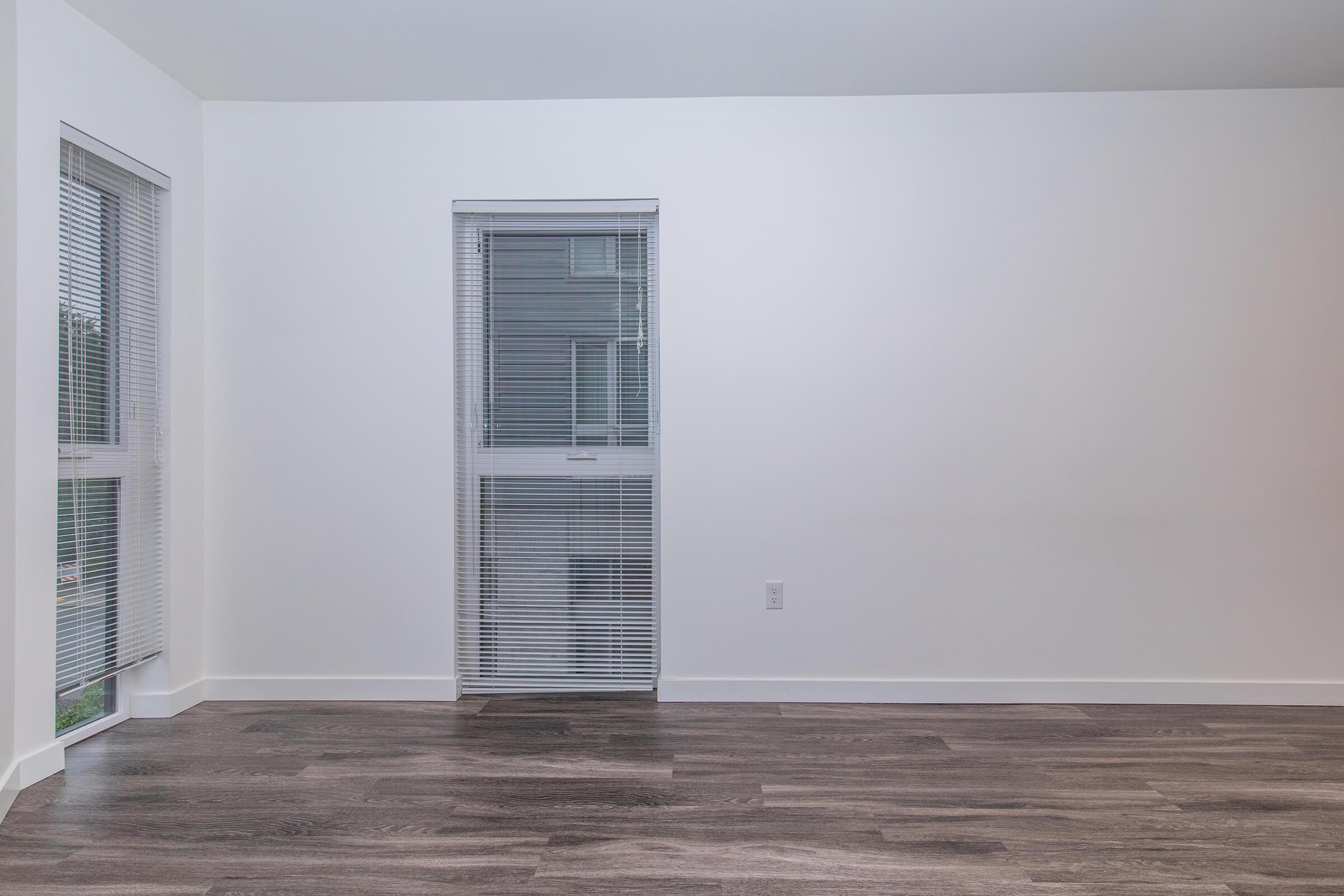 A bright, empty room with white walls and a large window featuring blinds. The flooring is dark wood. Natural light enters through the window, emphasizing the spaciousness of the room.