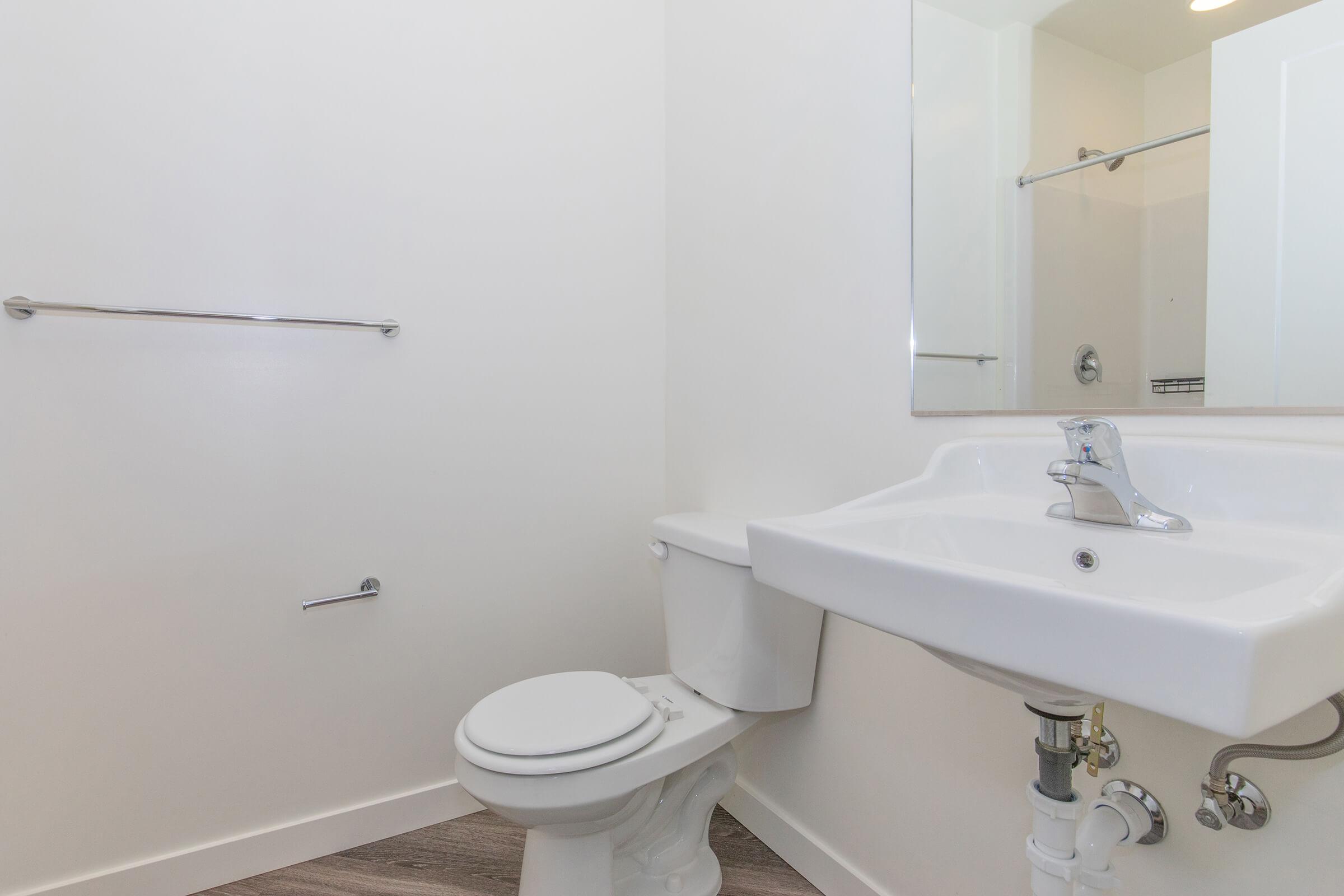 A clean, modern bathroom featuring a white toilet and a white sink with a sleek faucet. The walls are painted white, and there is a mirror above the sink. A towel bar is mounted on one wall, and the flooring is made of wood-like material, providing a bright and minimalist aesthetic.
