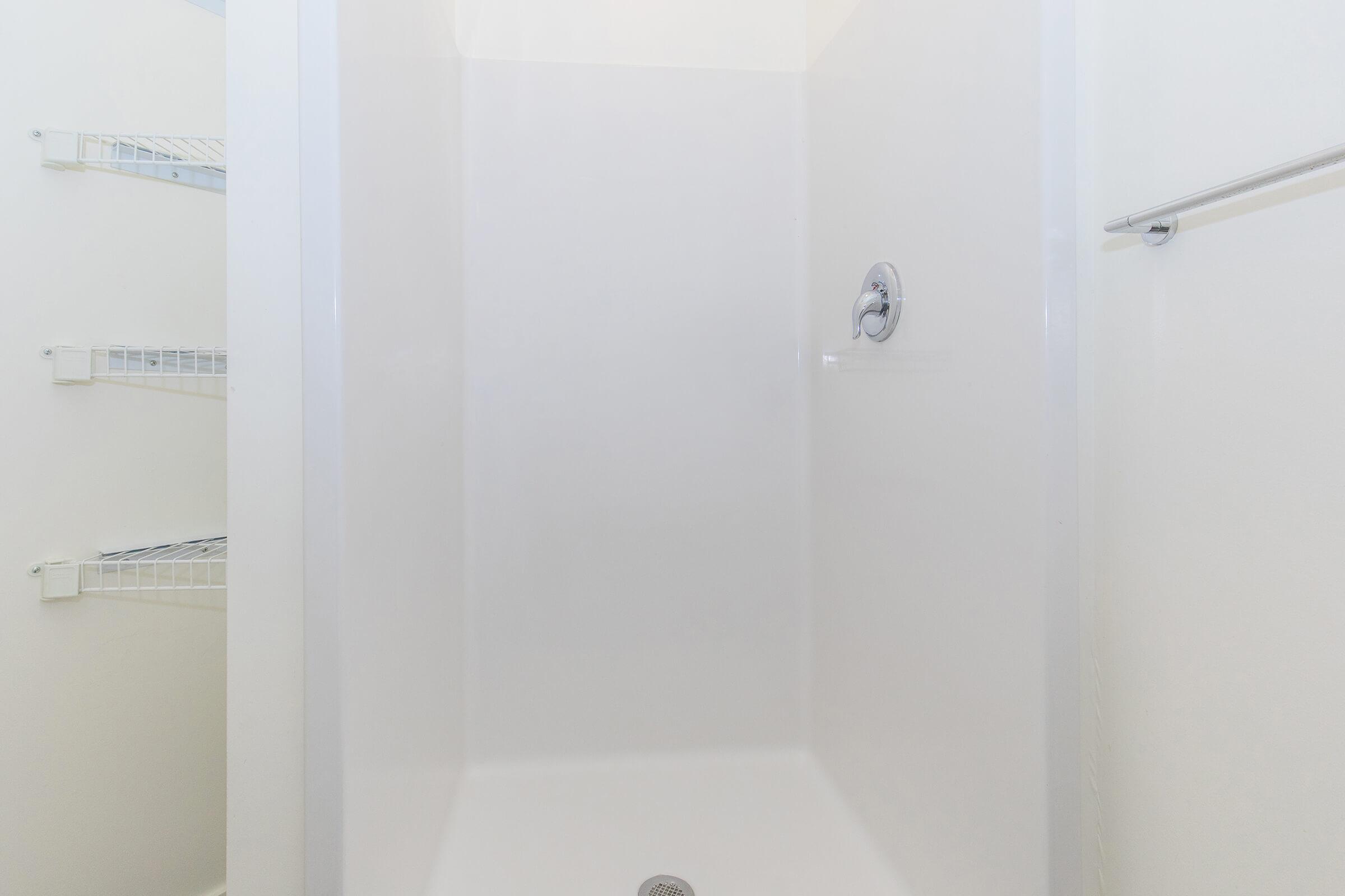 A clean, empty shower stall with a smooth, white finish. The walls are minimalistic, and there's a showerhead installed. In the background, there are metal wire shelves for storage. The overall space appears bright and well-maintained.