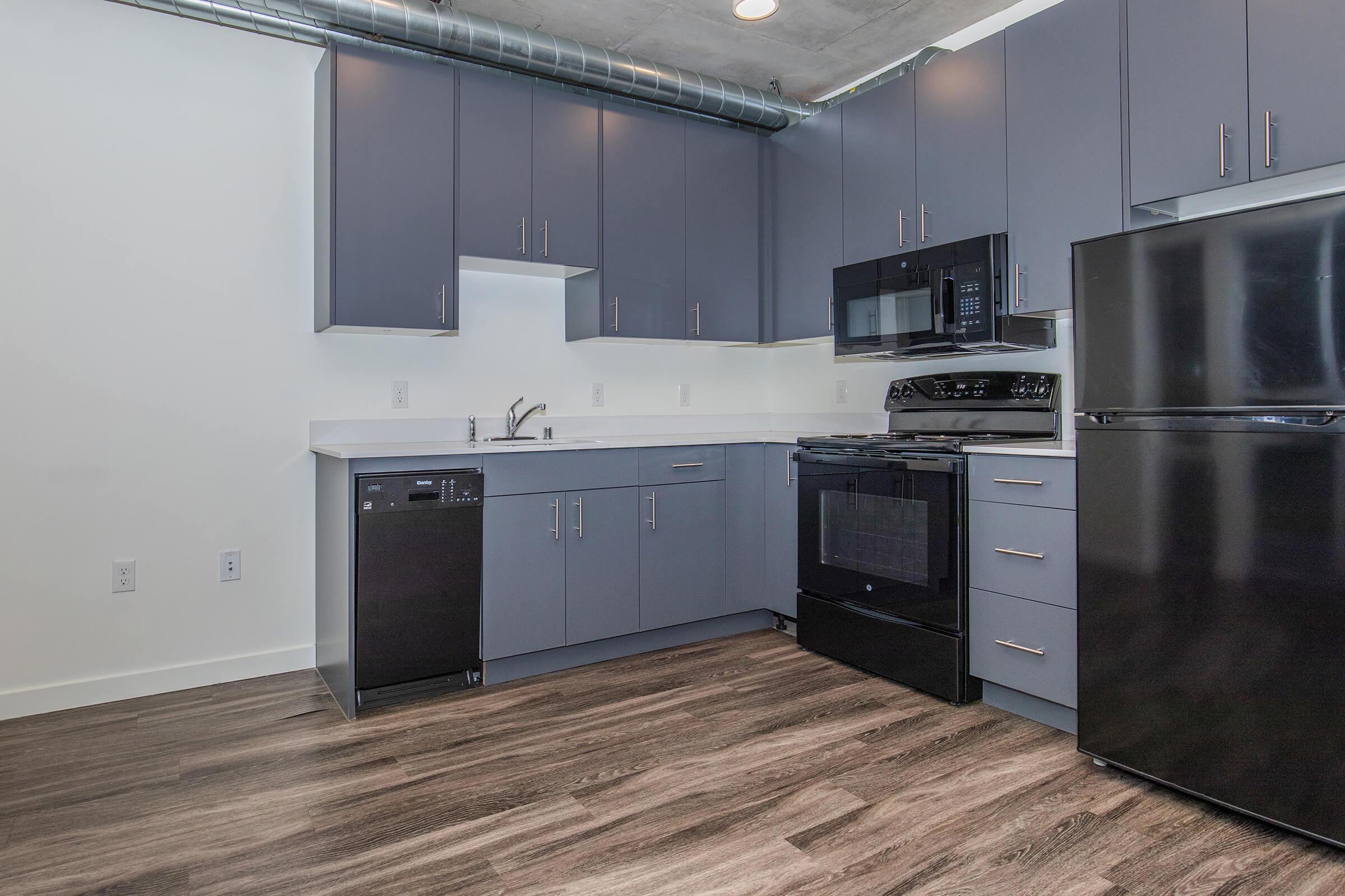 Modern kitchen featuring dark cabinetry, stainless steel appliances including a refrigerator, stove, microwave, and dishwasher. The countertops are light-colored, and the flooring is a wood-like laminate. Exposed ductwork runs along the ceiling, adding an industrial touch to the contemporary design.