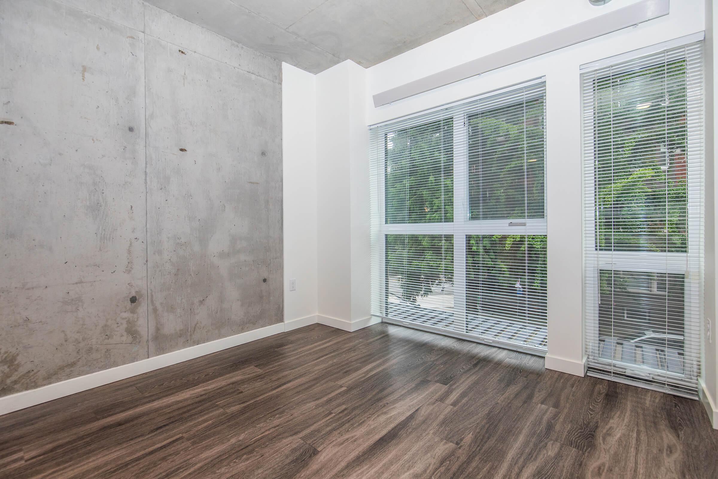 A modern, empty room featuring concrete walls and large windows with blinds. The floor is covered in dark wood laminate, and there is greenery visible outside through the windows. The design is minimalistic and contemporary, with a focus on natural light and open space.