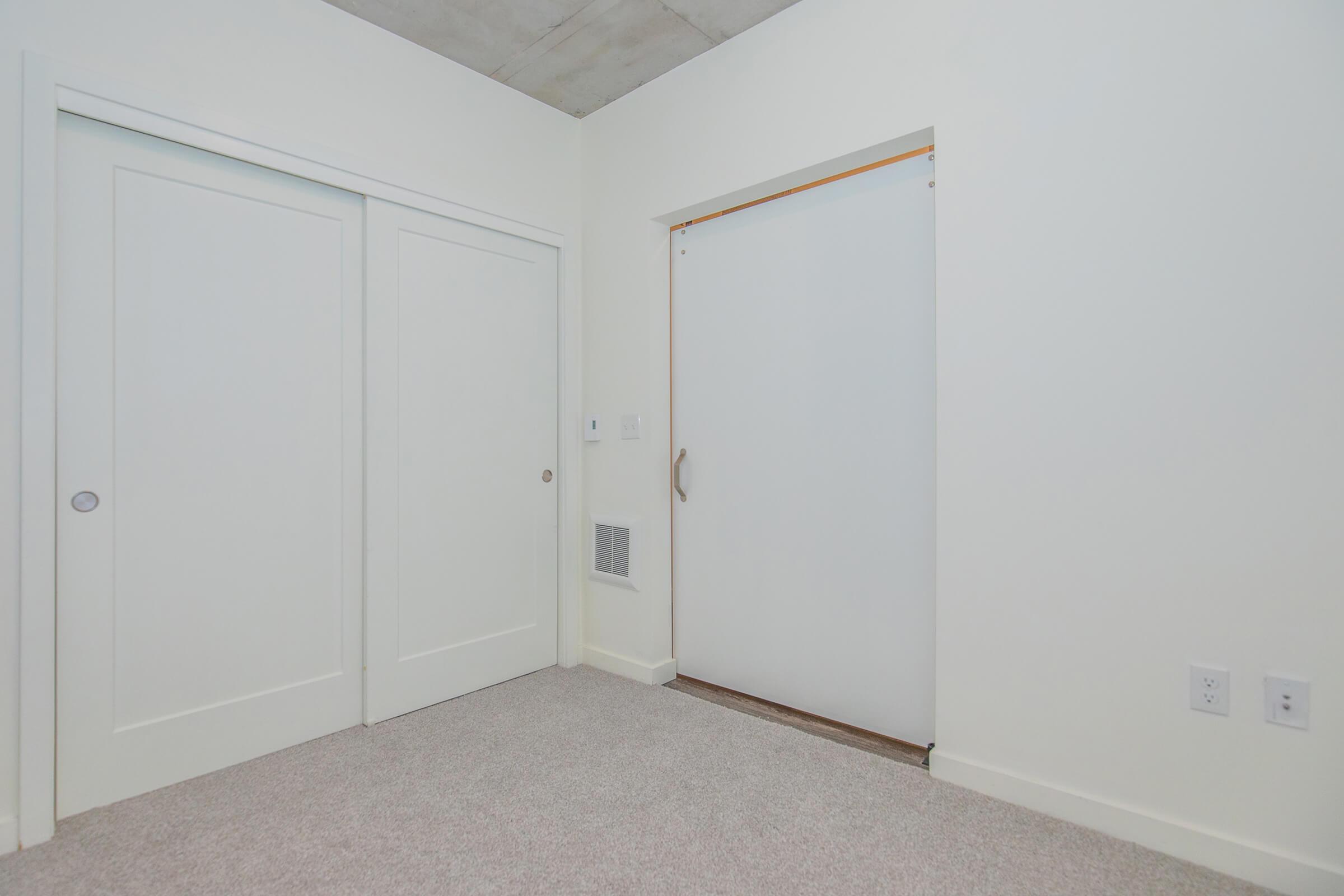 A simple, minimalist room featuring two white sliding closet doors and a sliding door on the right leading to another space. The walls are plain white, with light beige carpet flooring. The ceiling has a concrete finish. The room appears bright and uncluttered.