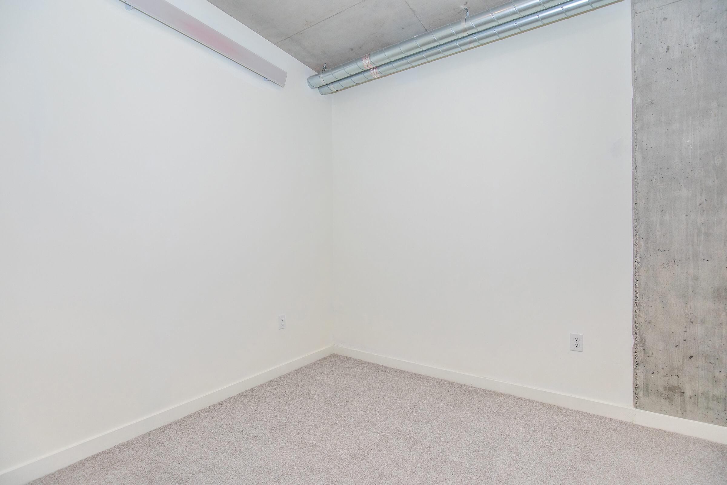A vacant room with light-colored walls, a concrete wall section, and a carpeted floor. The ceiling features exposed ductwork, and there are no furnishings or decorations present, creating a minimalist and empty atmosphere.