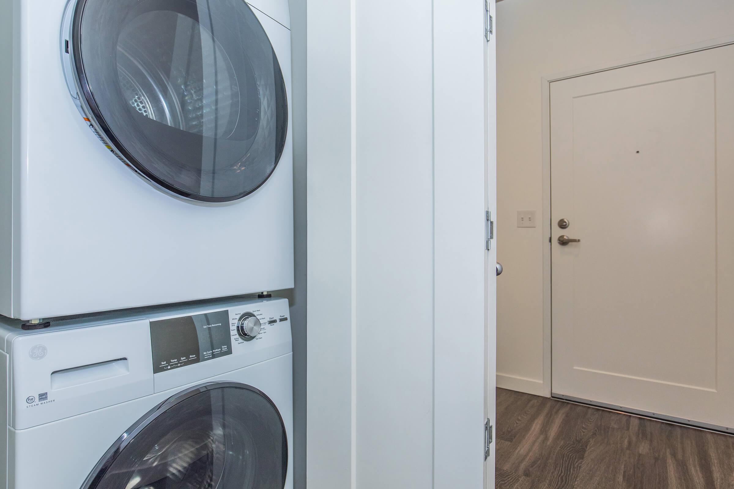 A modern laundry area featuring a stacked washing machine and dryer in sleek white design. There is a door leading outside visible in the background, and the floor is made of dark wood laminate. The space appears clean and well-organized.