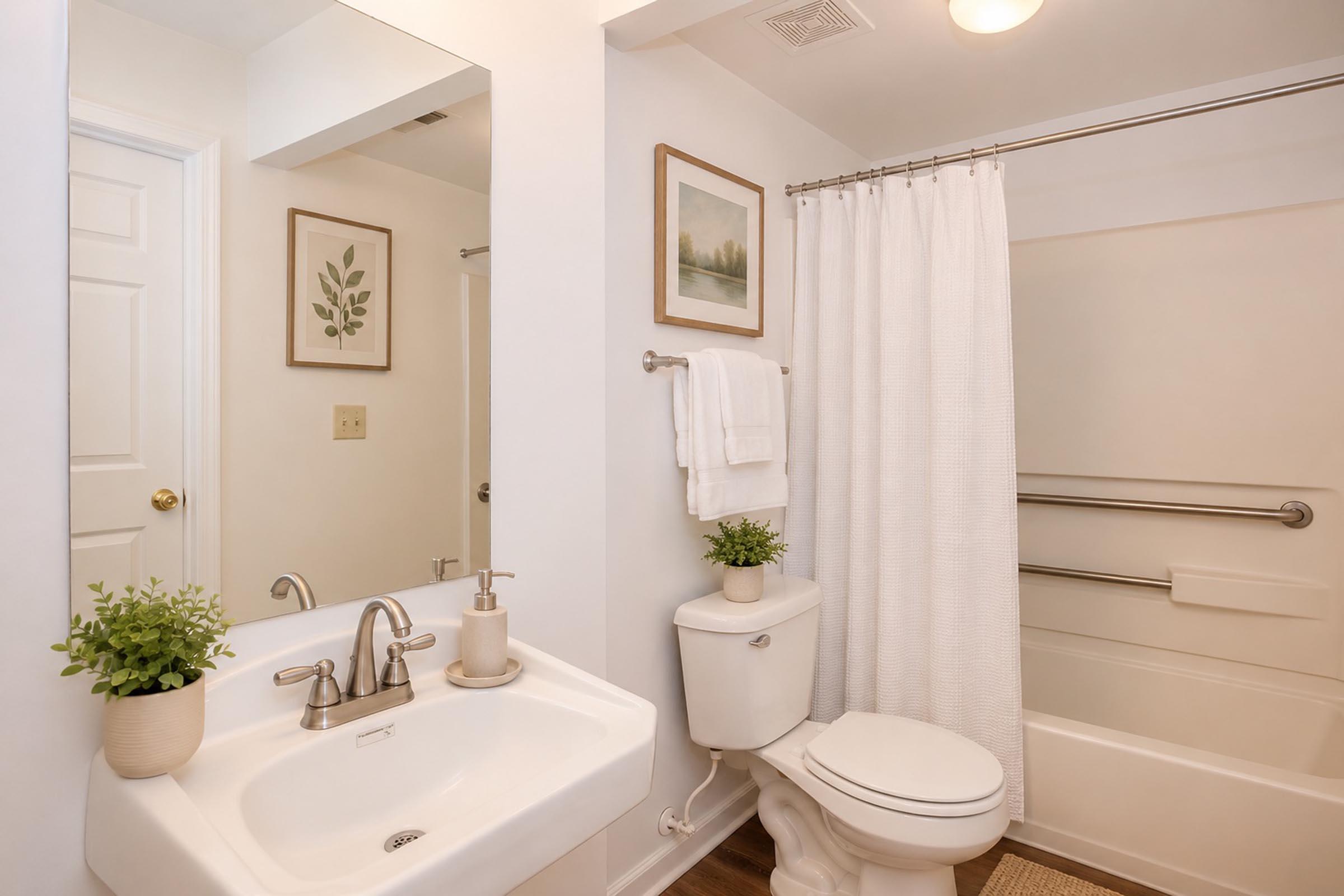 Bright and modern bathroom featuring a white sink with a stylish faucet, a white toilet, and a shower area with a white curtain. A small potted plant decorates the sink. The walls are adorned with a framed botanical print and a scenic landscape, creating a fresh and inviting atmosphere.