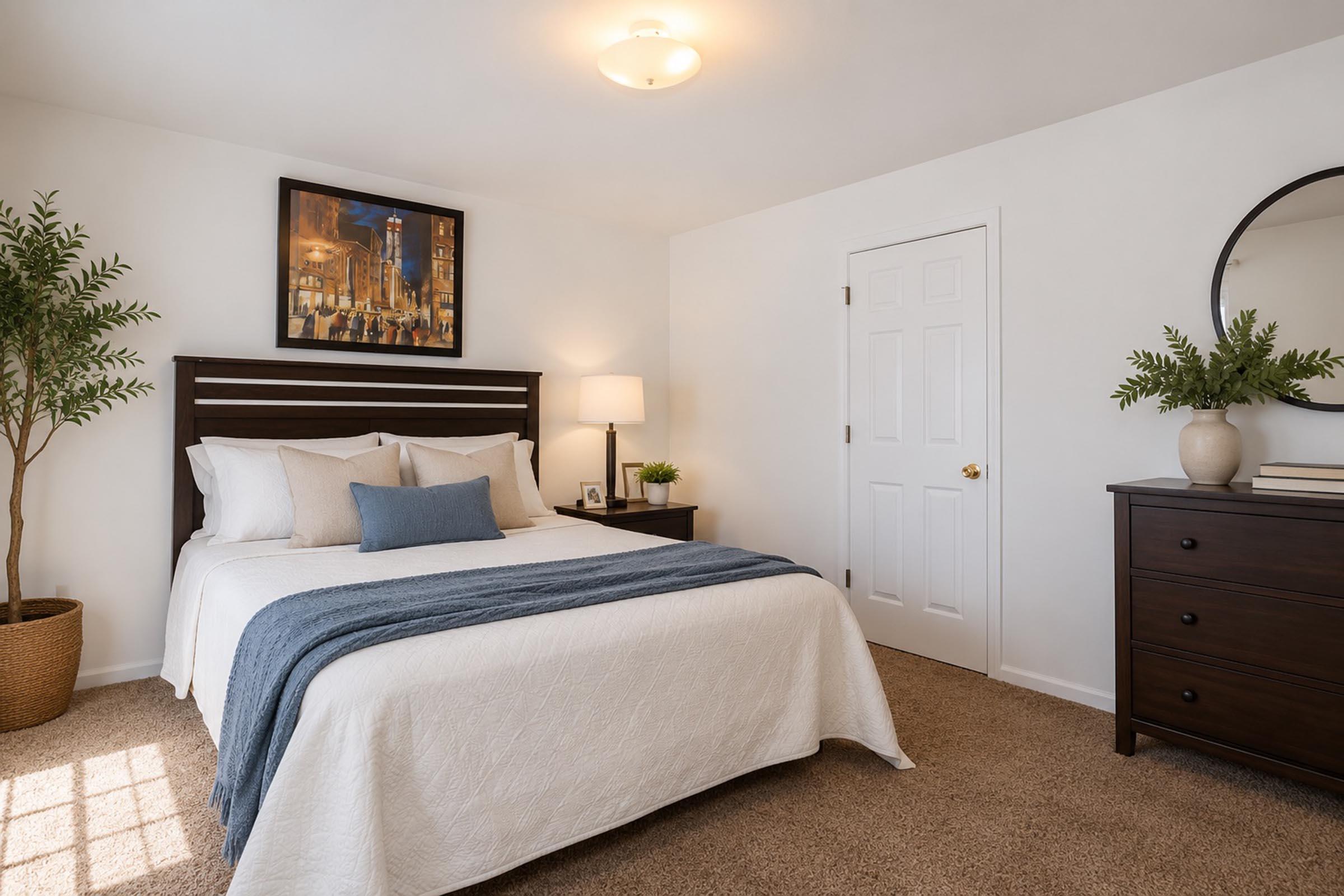 A cozy bedroom featuring a bed with a white bedspread and decorative cushions, a bedside lamp, and a dark wood dresser. A round mirror and a potted plant add to the decor. A piece of artwork hangs above the bed, and there’s a door leading to another room. The walls are painted white, creating a bright and inviting space.