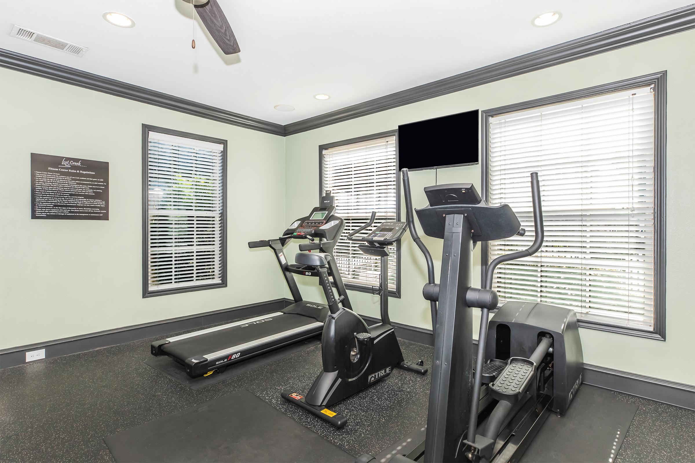 A well-lit fitness room featuring a treadmill and an elliptical machine. The walls are painted a soft green, and there are two large windows with blinds that allow natural light. A flat-screen TV is mounted on the wall, and there's a sign with information on the left side of the room.