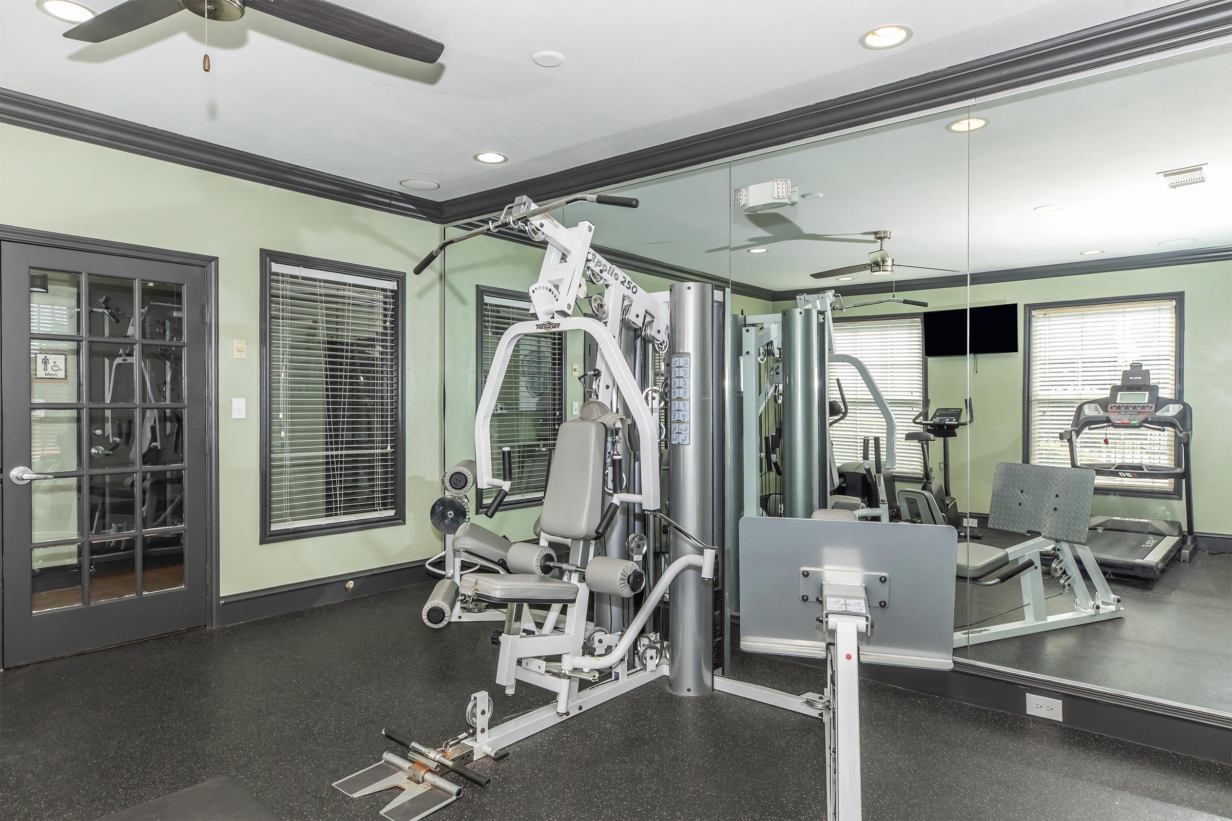 A modern fitness center featuring a multi-gym machine with a padded seat, surrounded by mirrors. In the background, additional workout equipment is visible, including cardio machines and weights. The walls are painted a soft green, and there are large windows providing natural light.