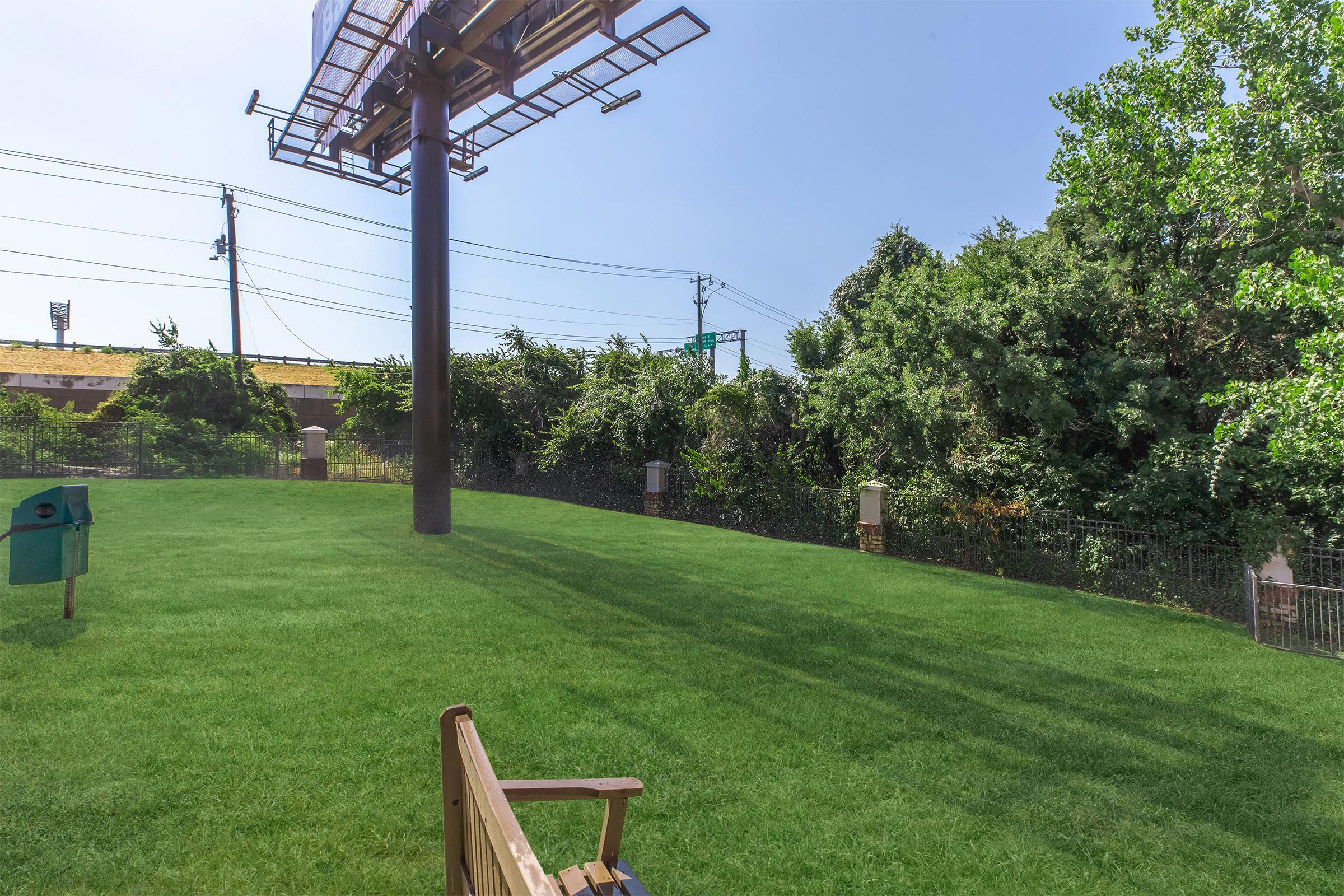 A grassy area with a wooden bench in the foreground, a large billboard supported by a pole behind it, and a lush green hedge in the background. There are utility poles and a few signs visible, creating a mix of nature and urban elements.