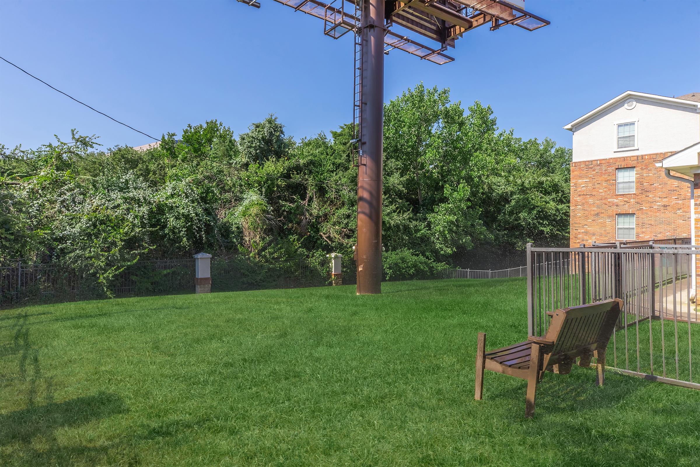 A grassy area in a backyard with a wooden bench, surrounded by trees and shrubs. In the background, there is a large metal pole and a brick building, along with a fenced section. The sky is clear and blue, creating a bright and open atmosphere.