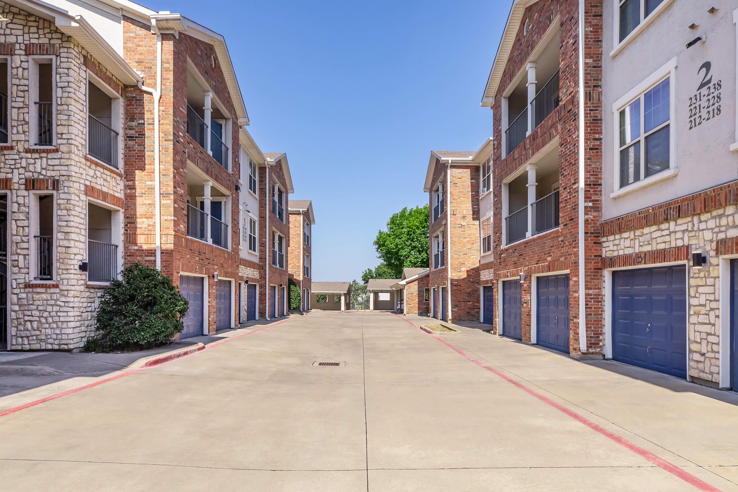 A residential complex with two lines of multi-story apartment buildings featuring brick and stone facades. The scene shows a paved driveway between the buildings, with garages visible. Clear blue sky overhead adds to the bright atmosphere of the area.