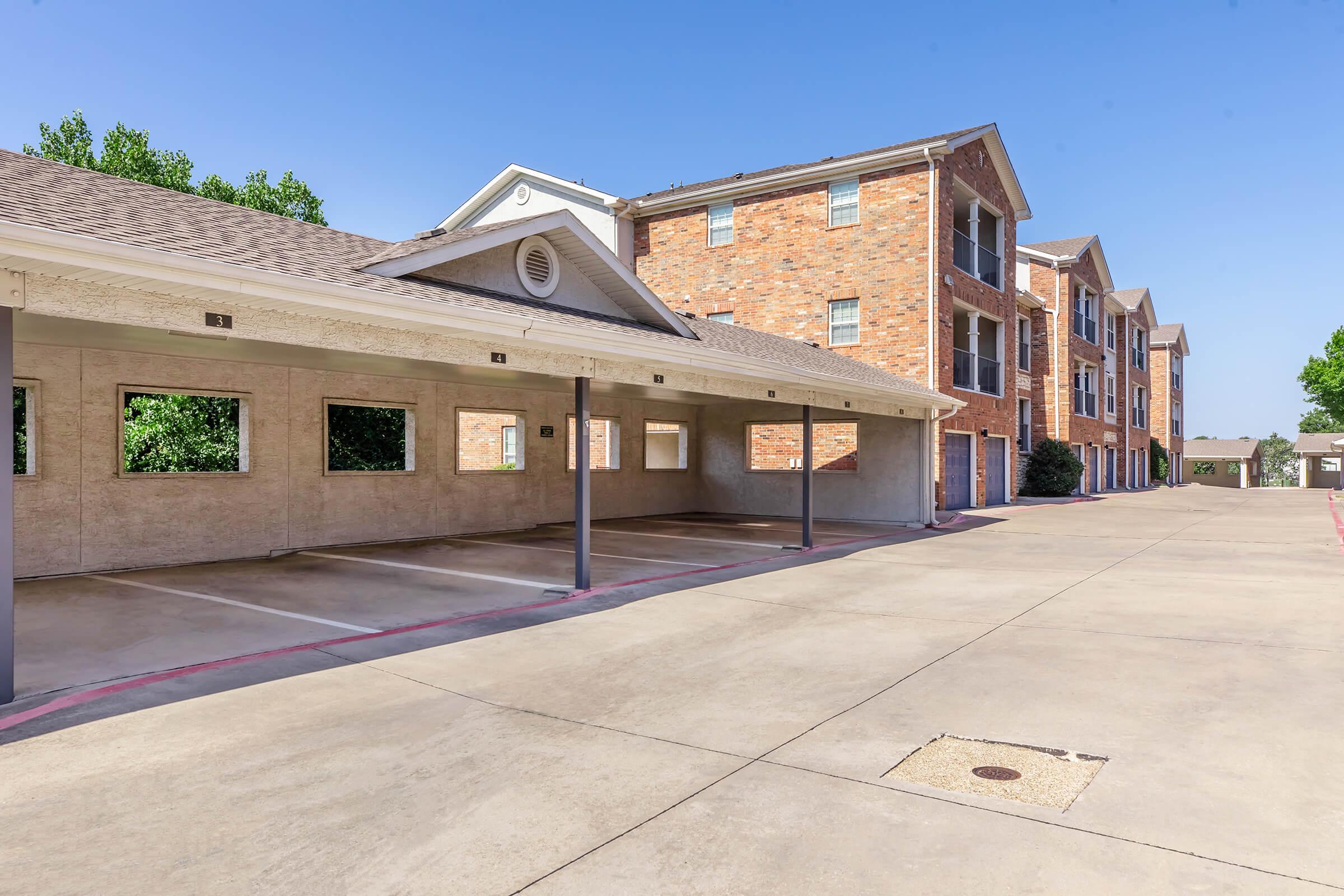 A parking area with covered spaces next to a three-story brick apartment building. The lot is clear with no vehicles parked. Surrounding greenery is visible, and the sky is clear and blue. The structure features multiple windows and a distinctive architectural design.