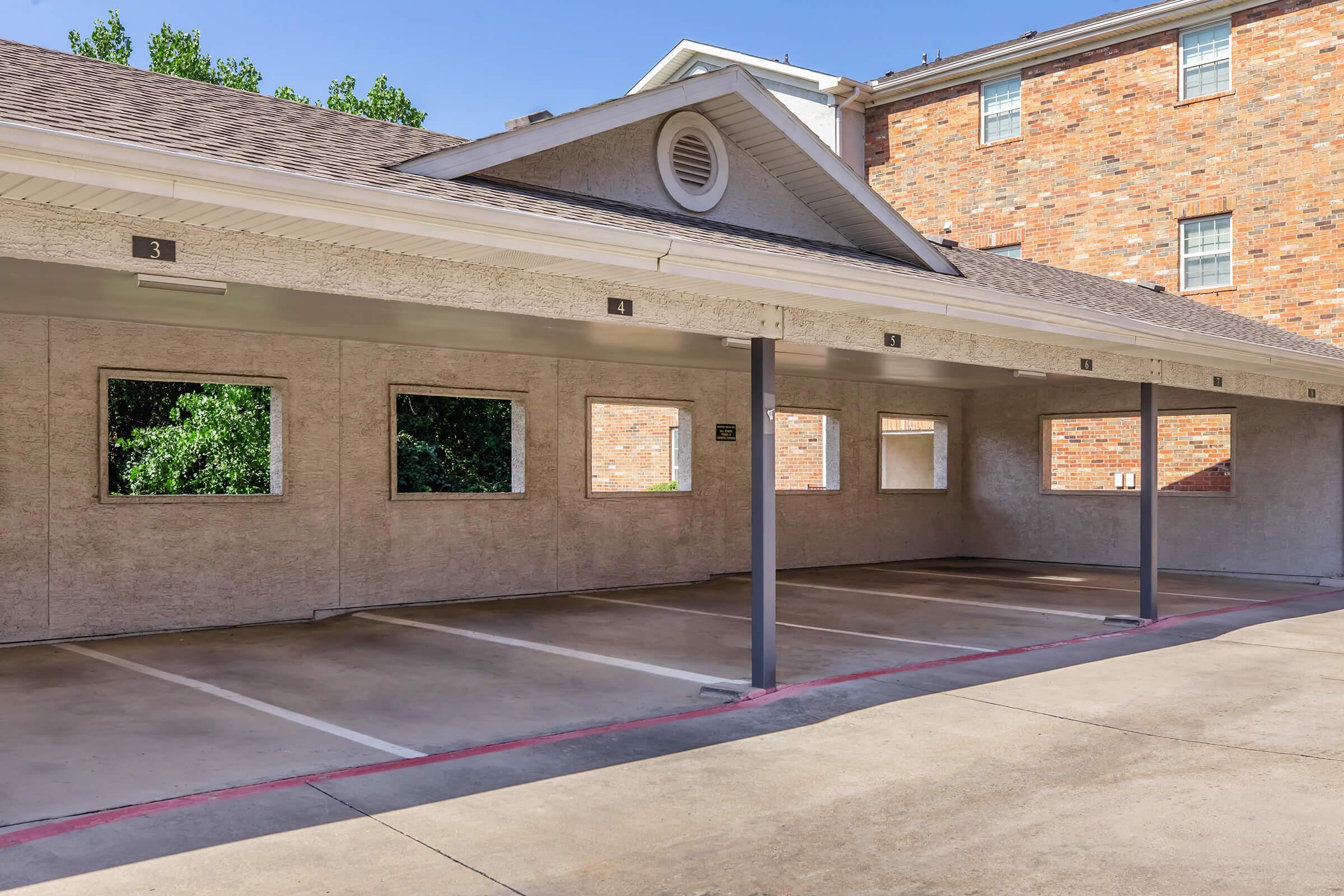 A vacant covered parking area with numbered spaces and several empty windows on the walls. The setting is within a residential building, featuring a brick exterior in the background and greenery visible outside the parking structure. The area is well-lit and clean.