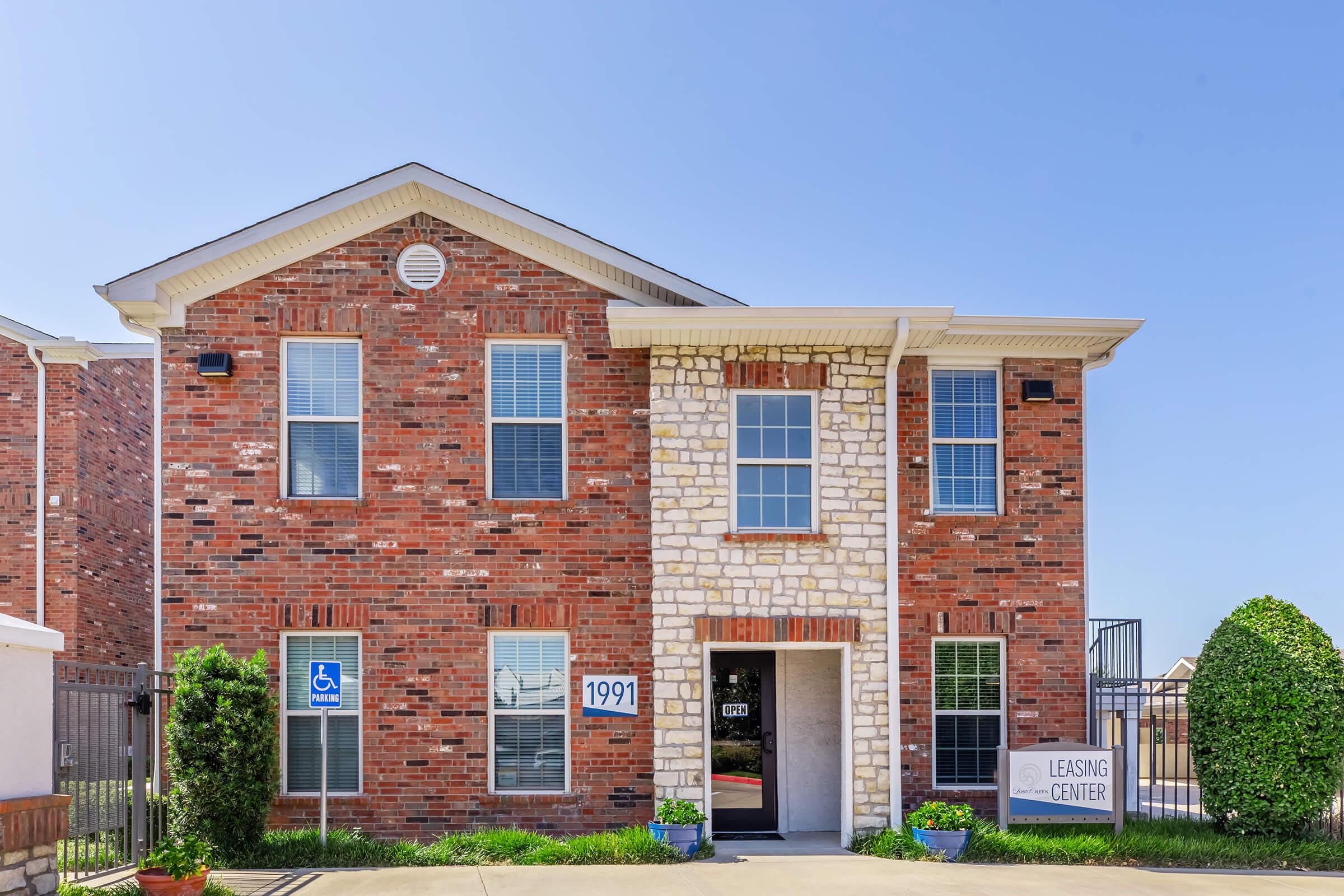 Image of a leasing center for an apartment complex. The building features a combination of red brick and stone exteriors, with several large windows. The entrance is marked by a sign that says "LEASING CENTER." There are landscaped green plants and a wheelchair accessible entrance. Clear blue sky overhead.