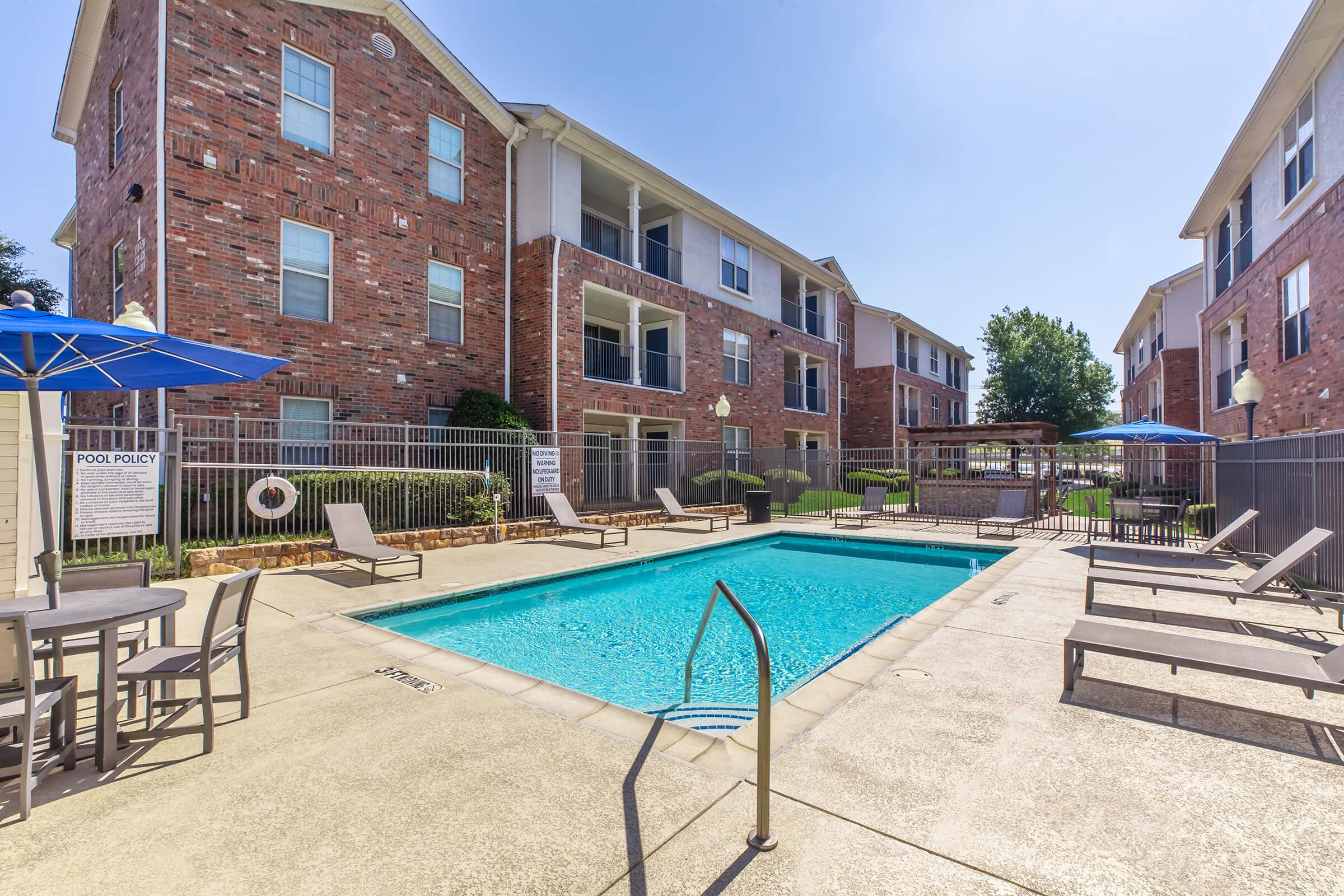 A bright outdoor pool area surrounded by brick apartment buildings. The pool is clear and inviting, with lounge chairs on the deck. There are umbrellas for shade and signs indicating pool policies. Green landscaping is visible in the background, creating a pleasant atmosphere.