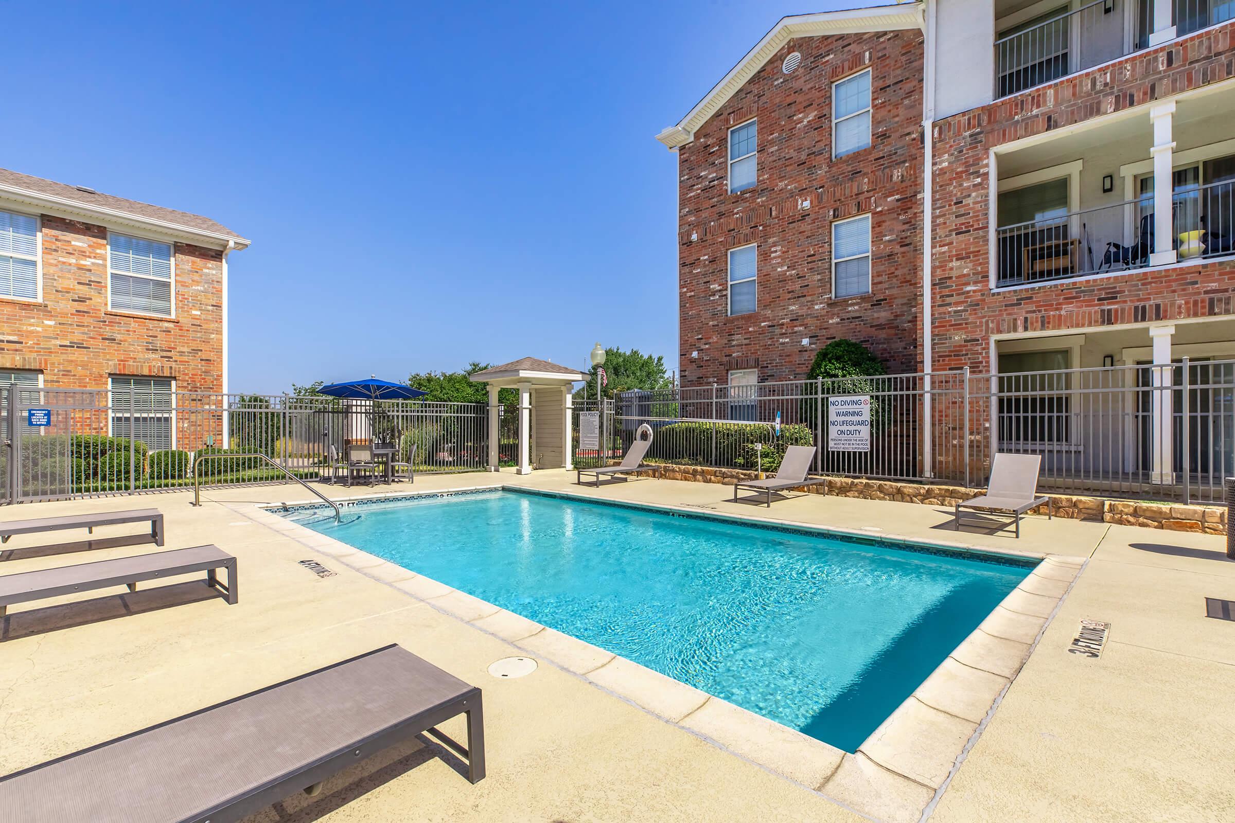 A clear blue swimming pool surrounded by lounge chairs, situated in a courtyard with brick buildings in the background. The area is enclosed by a fence, and there are umbrellas providing shade. The sky is bright and clear, indicating a sunny day.