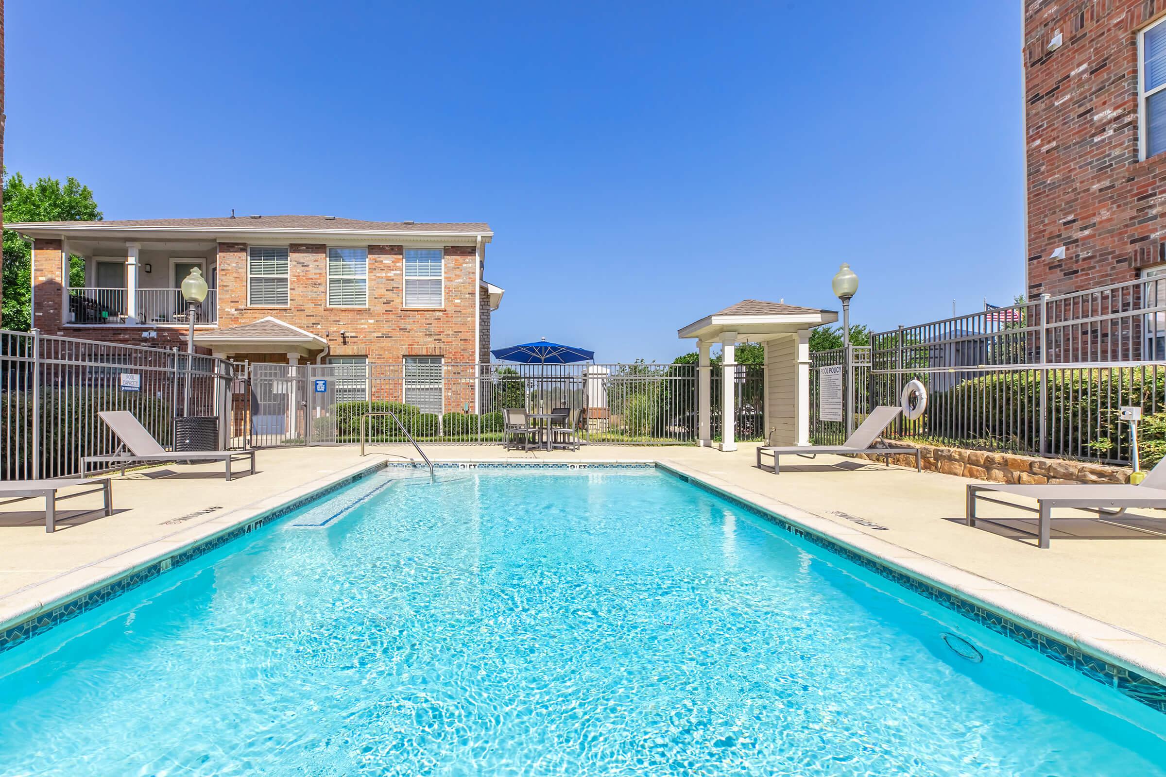A clear blue swimming pool surrounded by lounge chairs, with a fenced area and brick buildings in the background. The sky is bright and blue, creating a sunny atmosphere. This outdoor pool area is well-maintained, inviting relaxation and leisure.