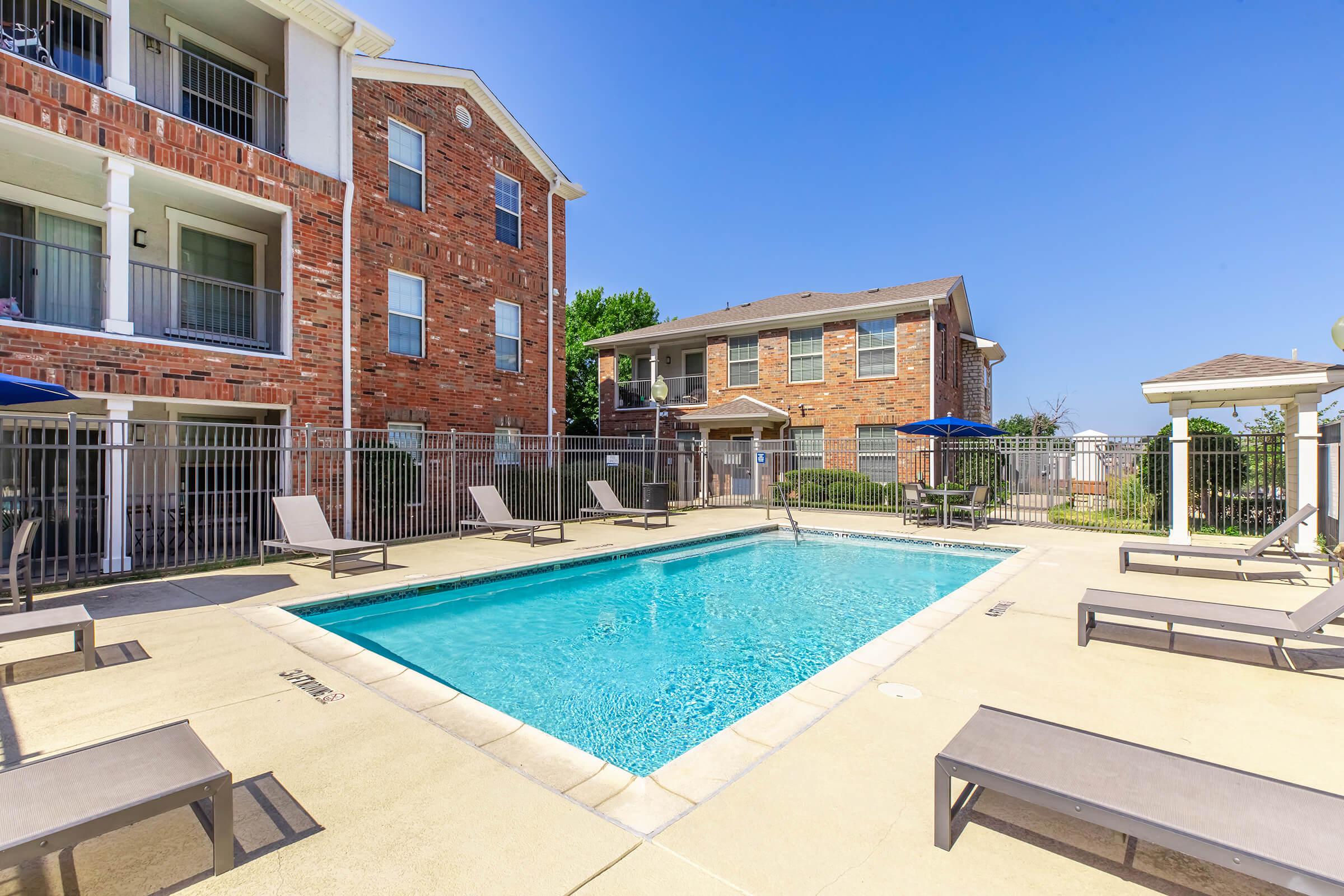 A clear blue swimming pool surrounded by lounge chairs in a residential area. Two multi-story brick buildings are visible in the background, along with shaded umbrellas and a well-maintained landscape. The sky is bright and sunny, creating a warm and inviting atmosphere.