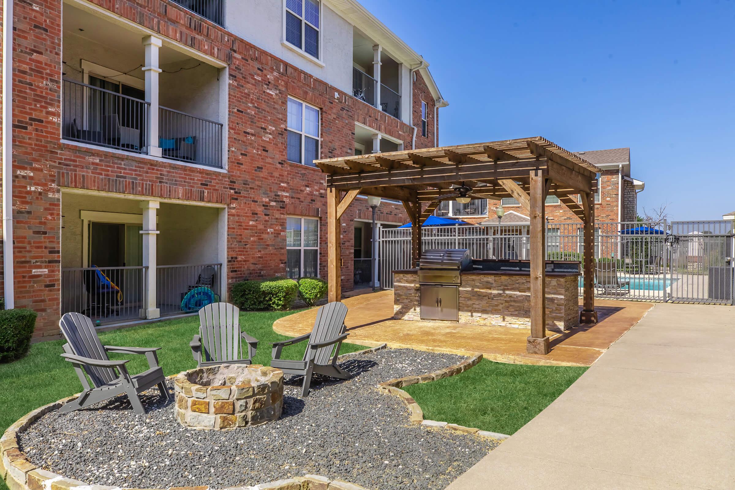 Outdoor area featuring a stone patio with a wooden pergola and a grill. There are several Adirondack chairs arranged around a fire pit, surrounded by landscaped grass and gravel. In the background, apartment buildings and a fenced swimming pool are visible under a clear blue sky.