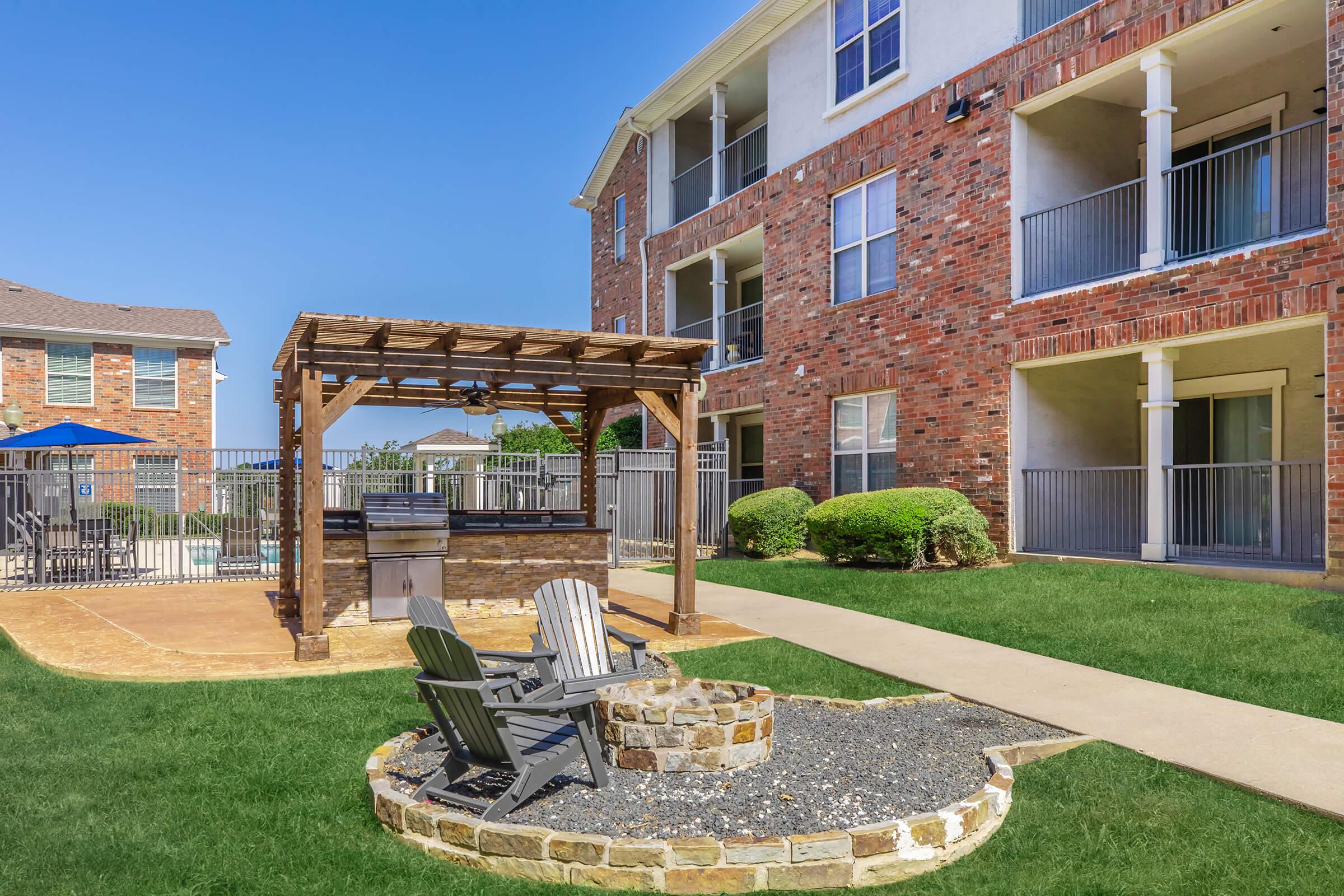 Outdoor area of an apartment complex featuring a stone fire pit surrounded by wooden chairs, a covered grill station, and well-manicured grass. In the background, there are two multi-story brick buildings with balconies, under a clear blue sky.