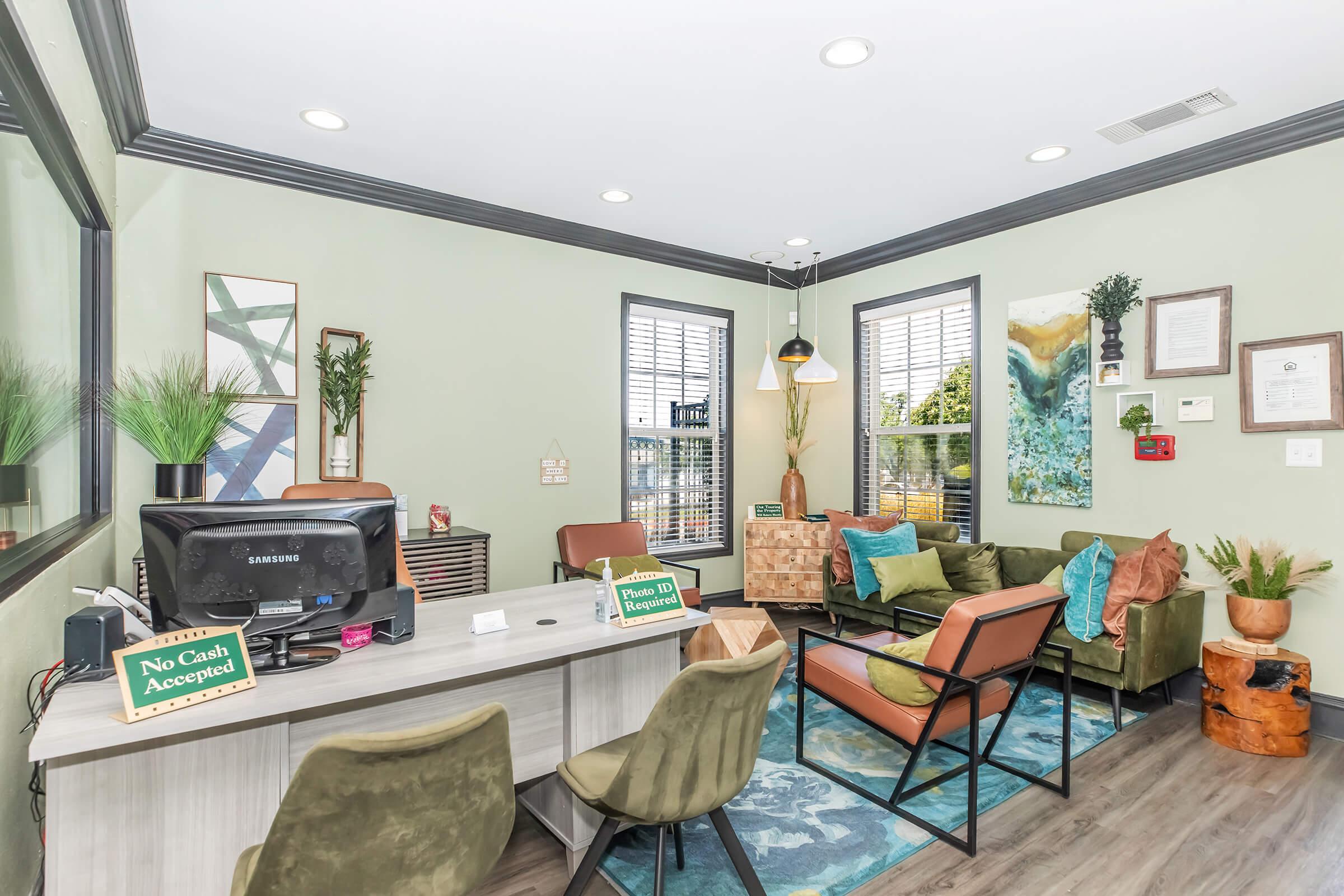 A cozy office reception area featuring comfortable seating, a computer desk, and decorative plants. The walls are painted in soft green, and there are large windows allowing natural light. Decorative art and a small wooden table enhance the inviting atmosphere, alongside signs indicating payment policies.