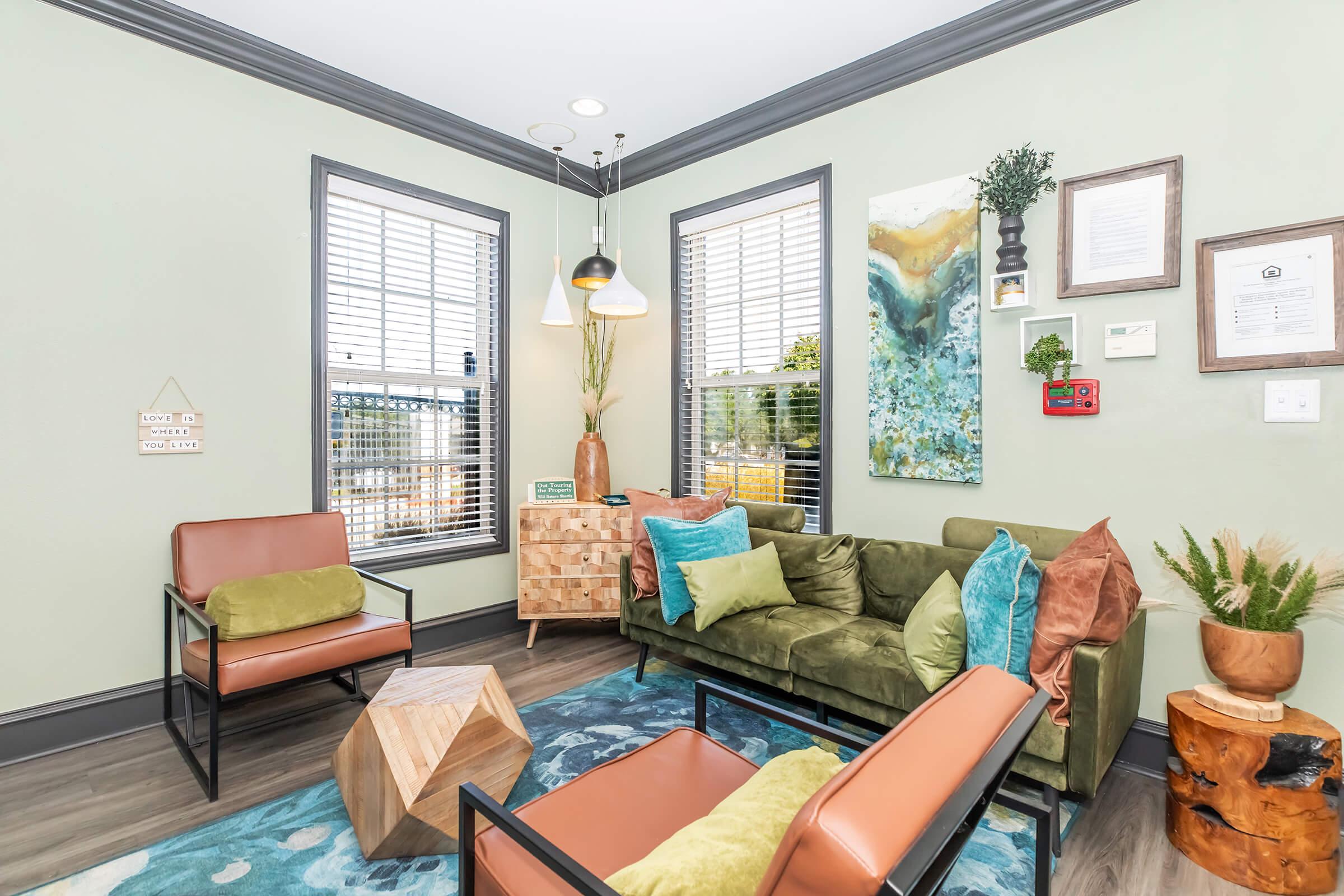 A cozy and modern lounge area featuring a green velvet sofa with colorful cushions, two stylish armchairs, a wooden coffee table, and decorative plants. Large windows let in natural light, while art and framed certificates adorn the walls in soft green tones. The space is inviting and well-decorated.