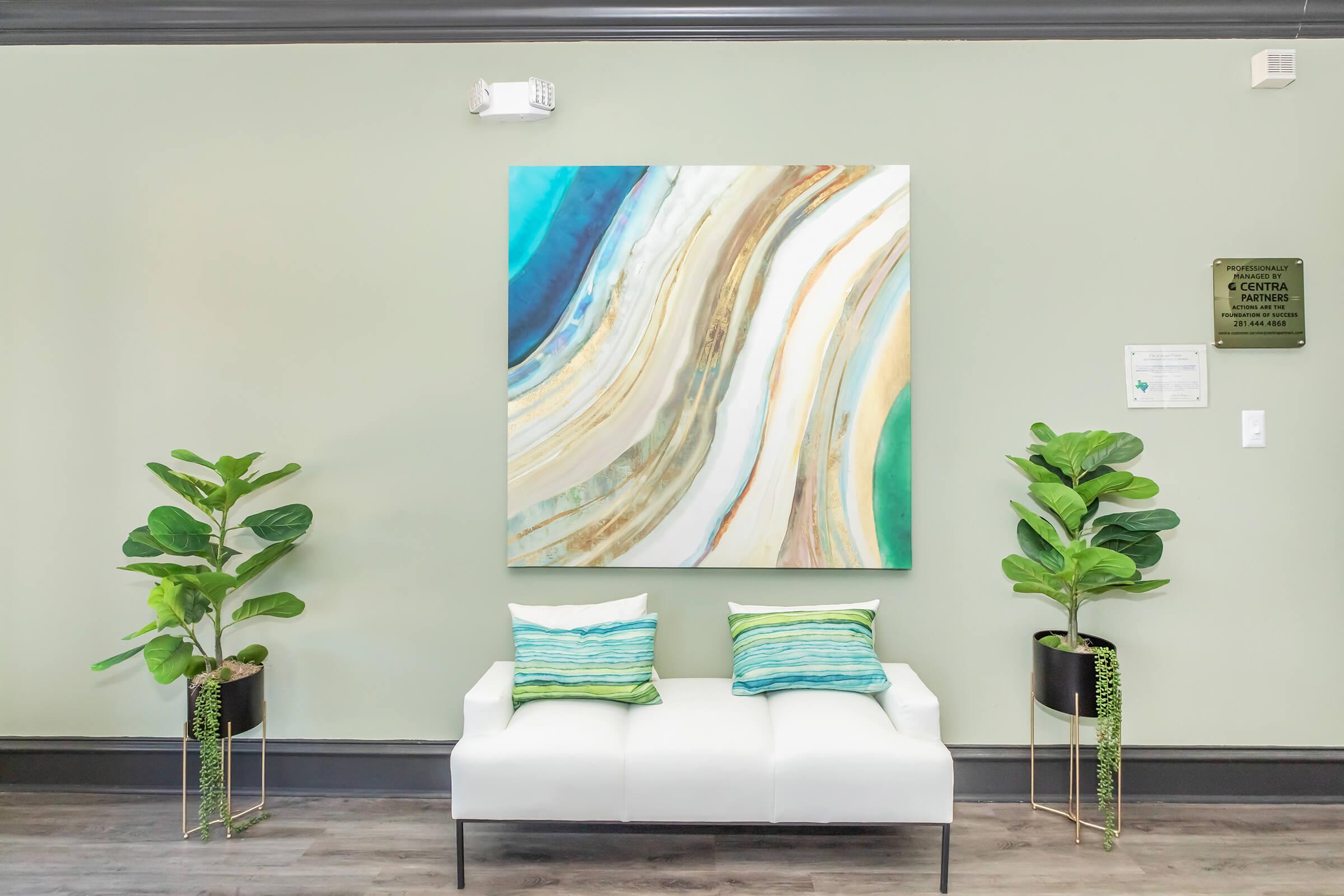 A modern waiting area featuring a white couch with green and blue decorative pillows. Two tall potted plants flank the couch. Behind the couch is a large abstract painting with wavy patterns in shades of teal, gold, and cream. The walls are painted a soft green, creating a calming atmosphere.