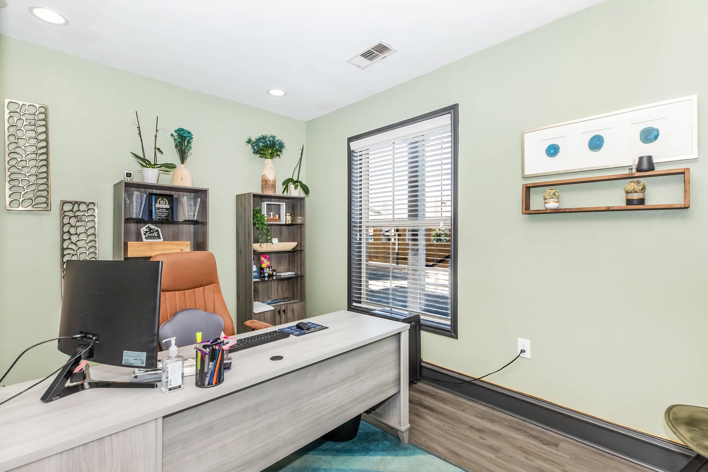 Modern office space featuring a wooden desk with an orange chair, a computer monitor, and various office supplies. The walls are painted a soft green, adorned with decorative shelves and plants. A window allows natural light in, creating a bright and inviting atmosphere.